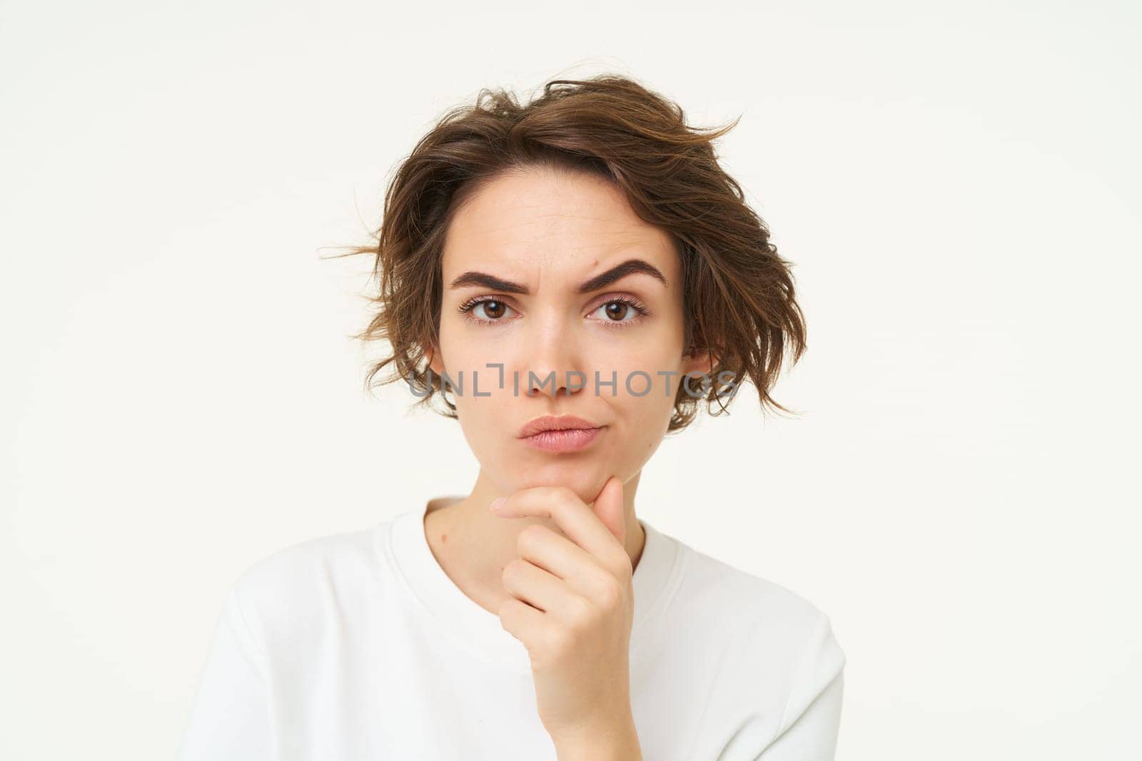 Portrait of thinking woman, touches her chin, looks serious and thoughtful, poses over white background. Copy space