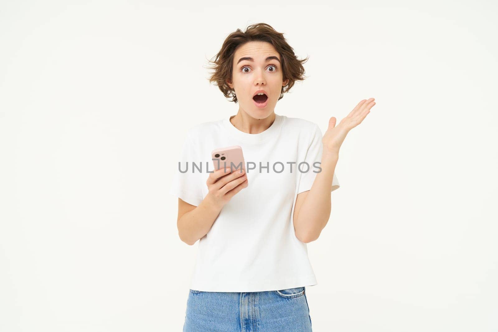 Portrait of brunette girl with shocked face, reading something amazing on mobile phone, standing with smartphone over white background.