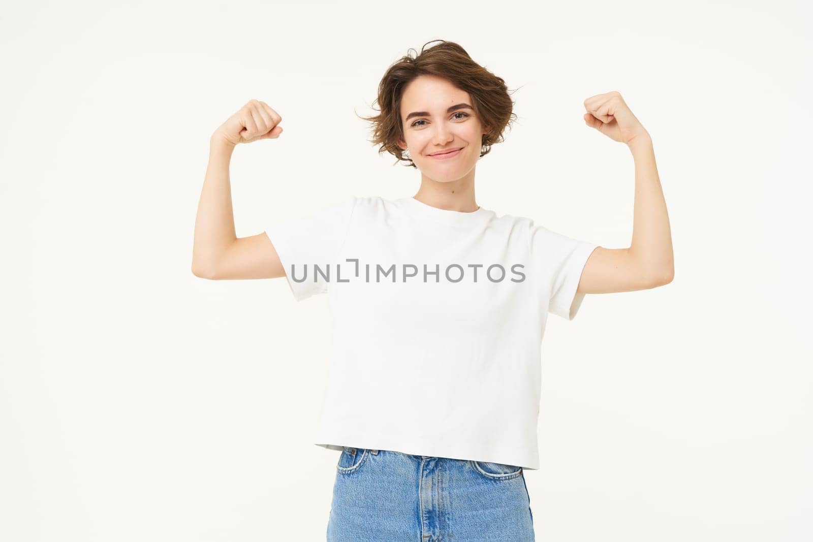 Portrait of confident and strong young woman, flexing biceps, shows her muscles on arms with proud smiling face, posing over white background.