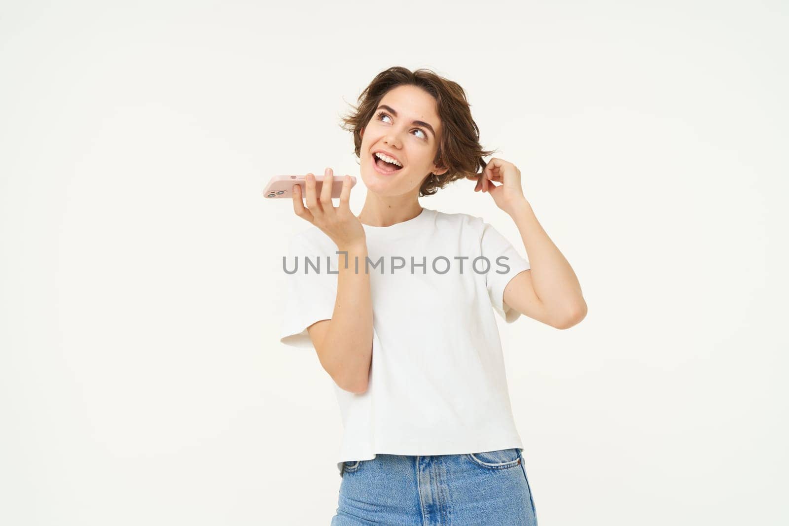 Portrait of chatty woman talking in speakerphone, records voice message on mobile phone and smiling, stands over white background.