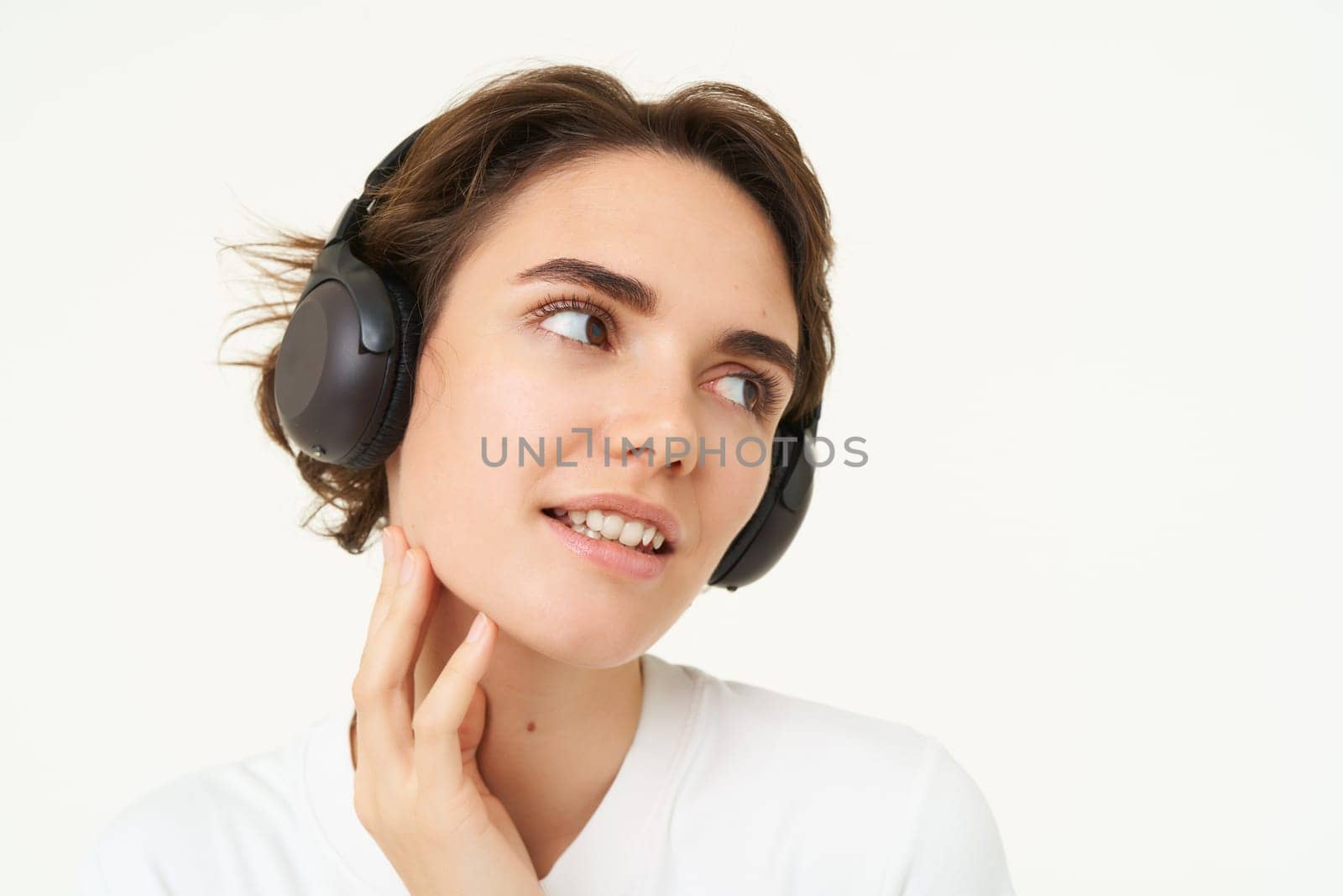 Image of young modern woman in wireless headphones, using earphones to listen to music, standing over white background. Copy space
