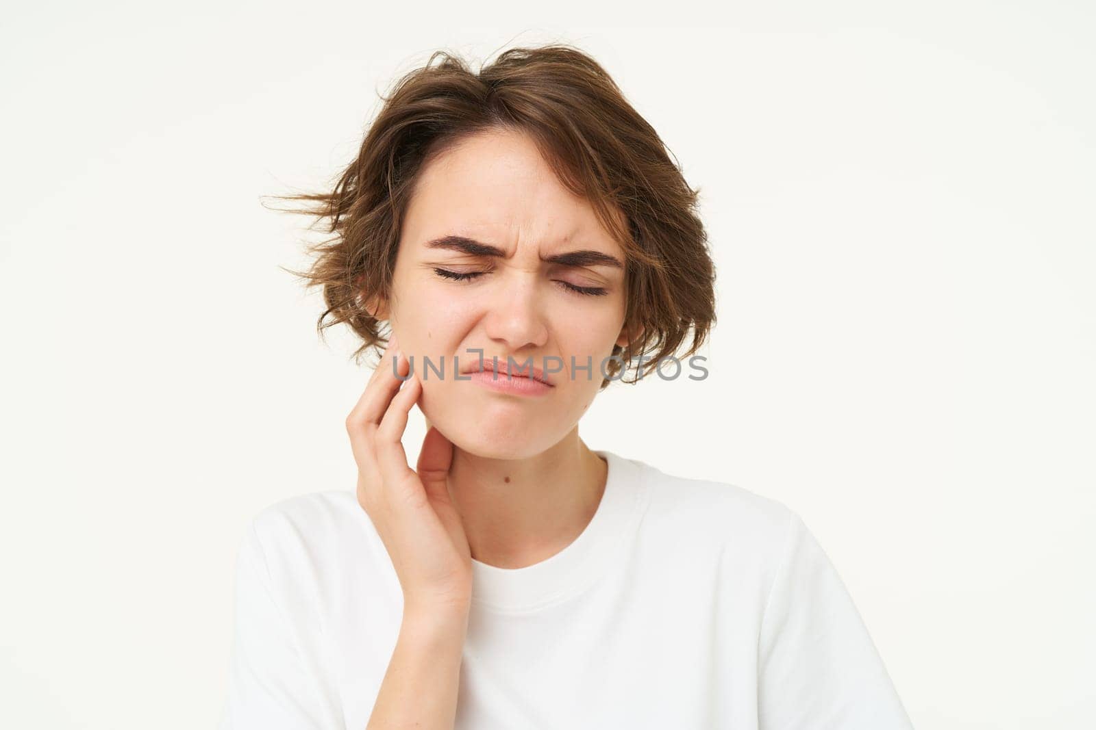 Close up of woman has a toothache, touches her teeth and frowns from painful discomfort, stands over white background by Benzoix