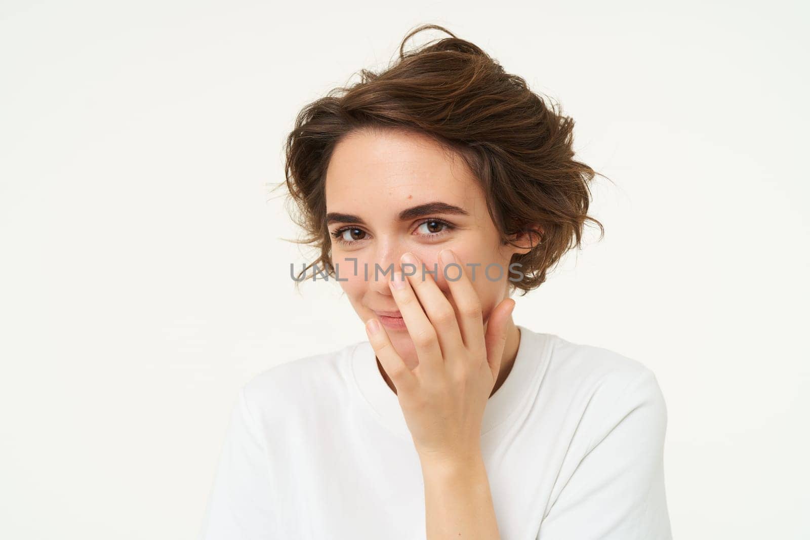 Close up of cute young brunette woman, covers her face, laughs and smiles, looks shy and silly, giggle, stands over white background.