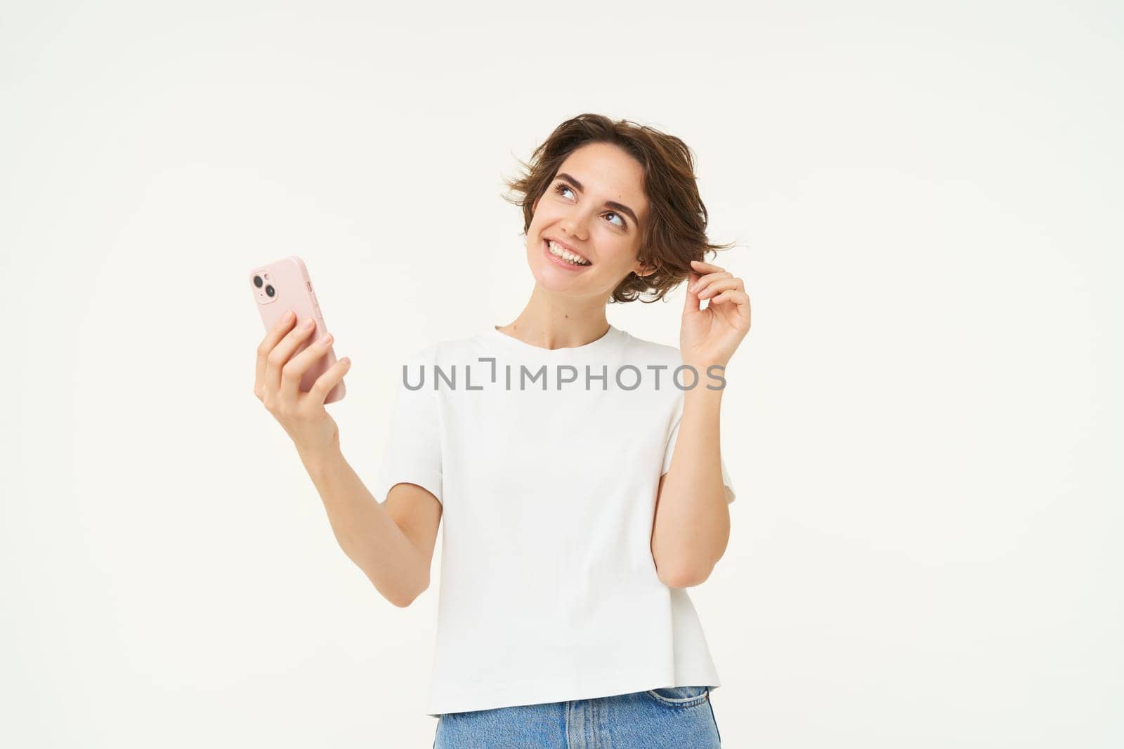 Portrait of happy brunette girl with smartphone, thinking with coquettish smile, standing over white background.