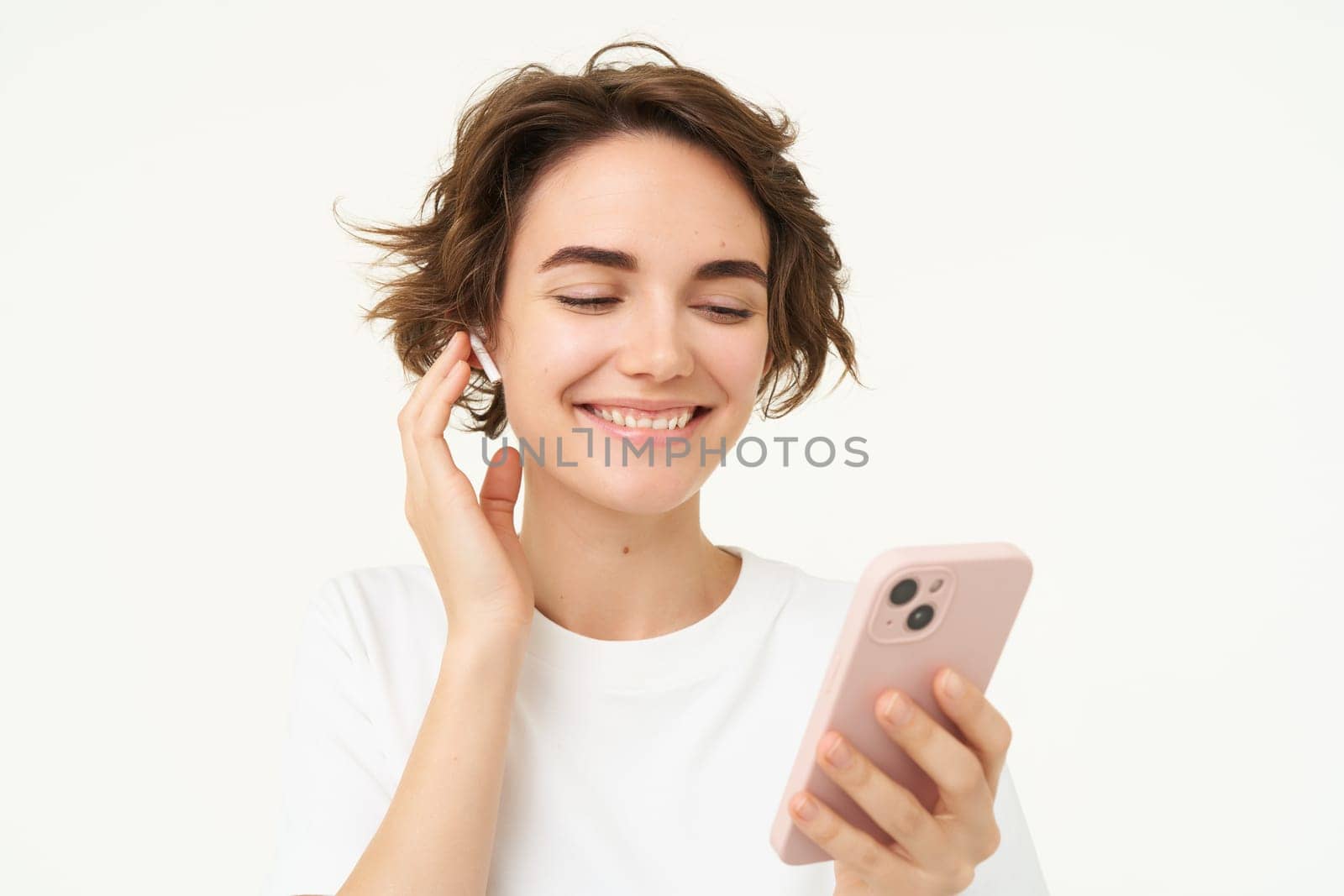 Woman singing along, listening music in wireless headphones, holding smartphone in hand, standing over white background.