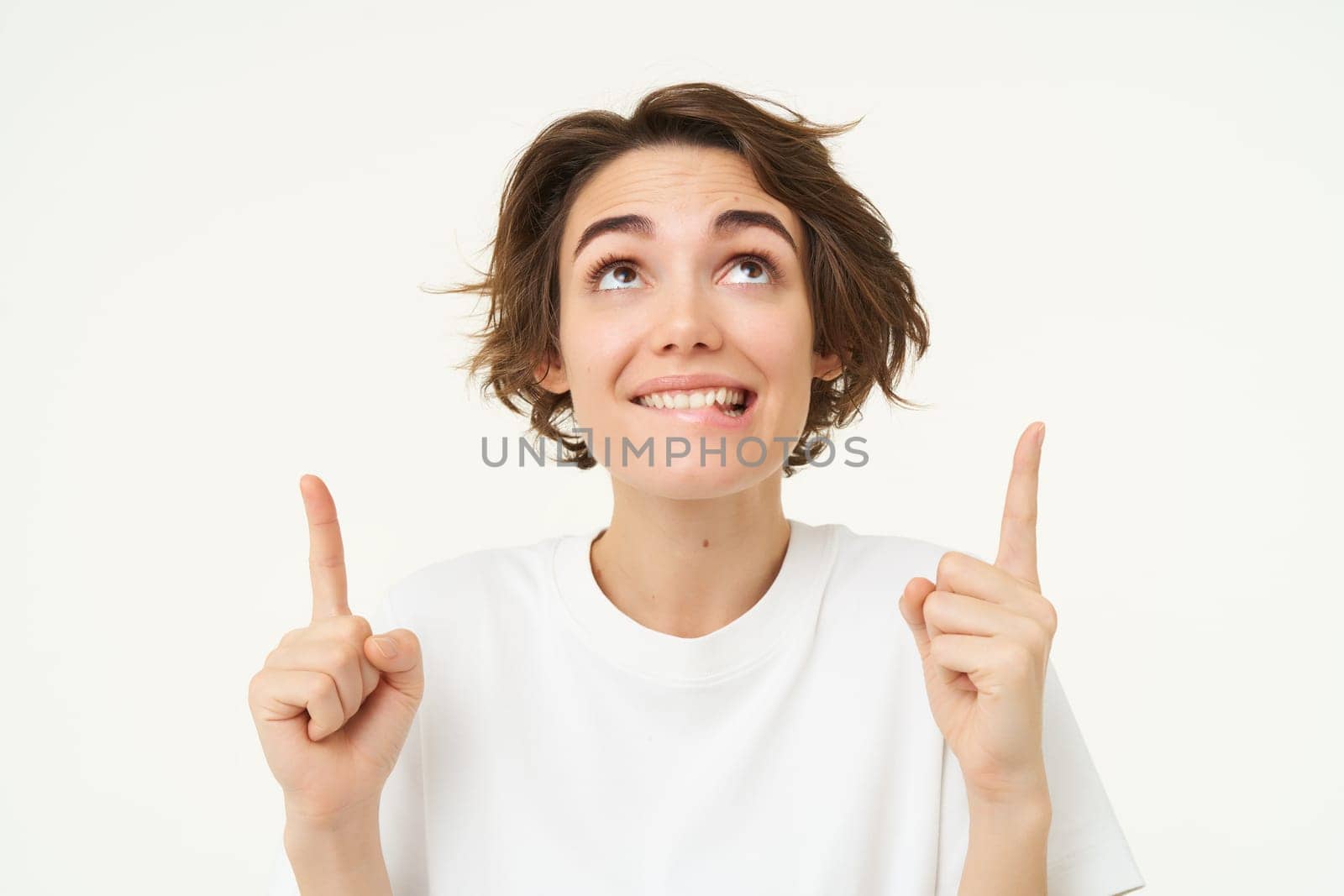 Portrait of happy, beautiful young woman pointing fingers up, showing advertisement or product on top of copy space, standing against white background by Benzoix