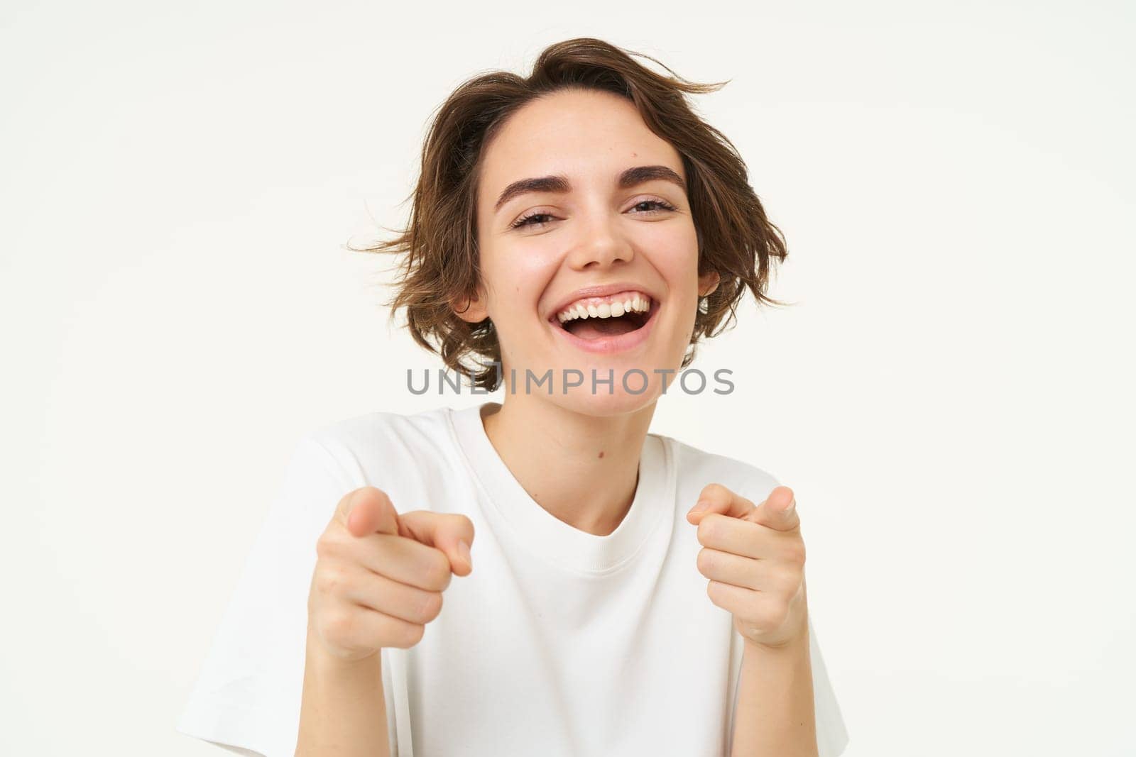 Close up portrait of cute smiling woman, pointing fingers at camera, choosing, inviting you, posing against white studio background.