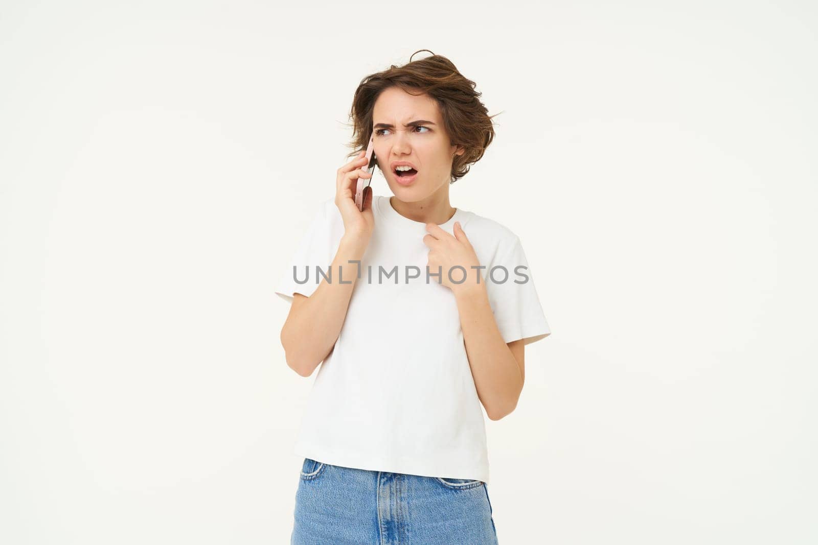 Portrait of shocked, angry woman talks on mobile phone with disappointed, furious face expression, standing over white background.