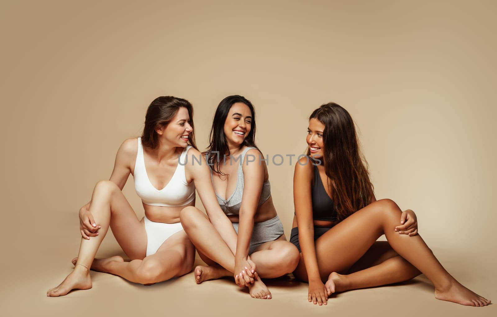 Cheerful millennial international women in underwear sit on floor, enjoy self love, lifestyle, isolated on beige background, studio, full length. Motivation, beauty care treatments and wellness