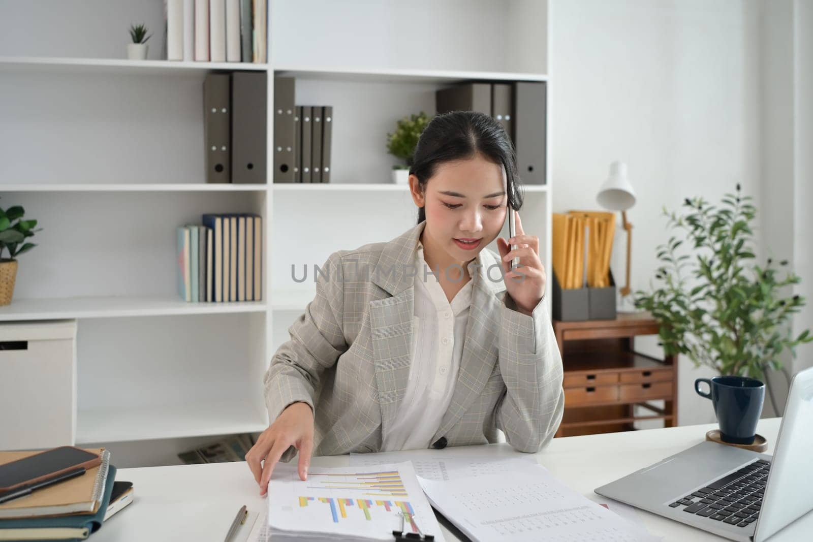 Smiling female accountant talking on mobile phone and checking financial document at office.