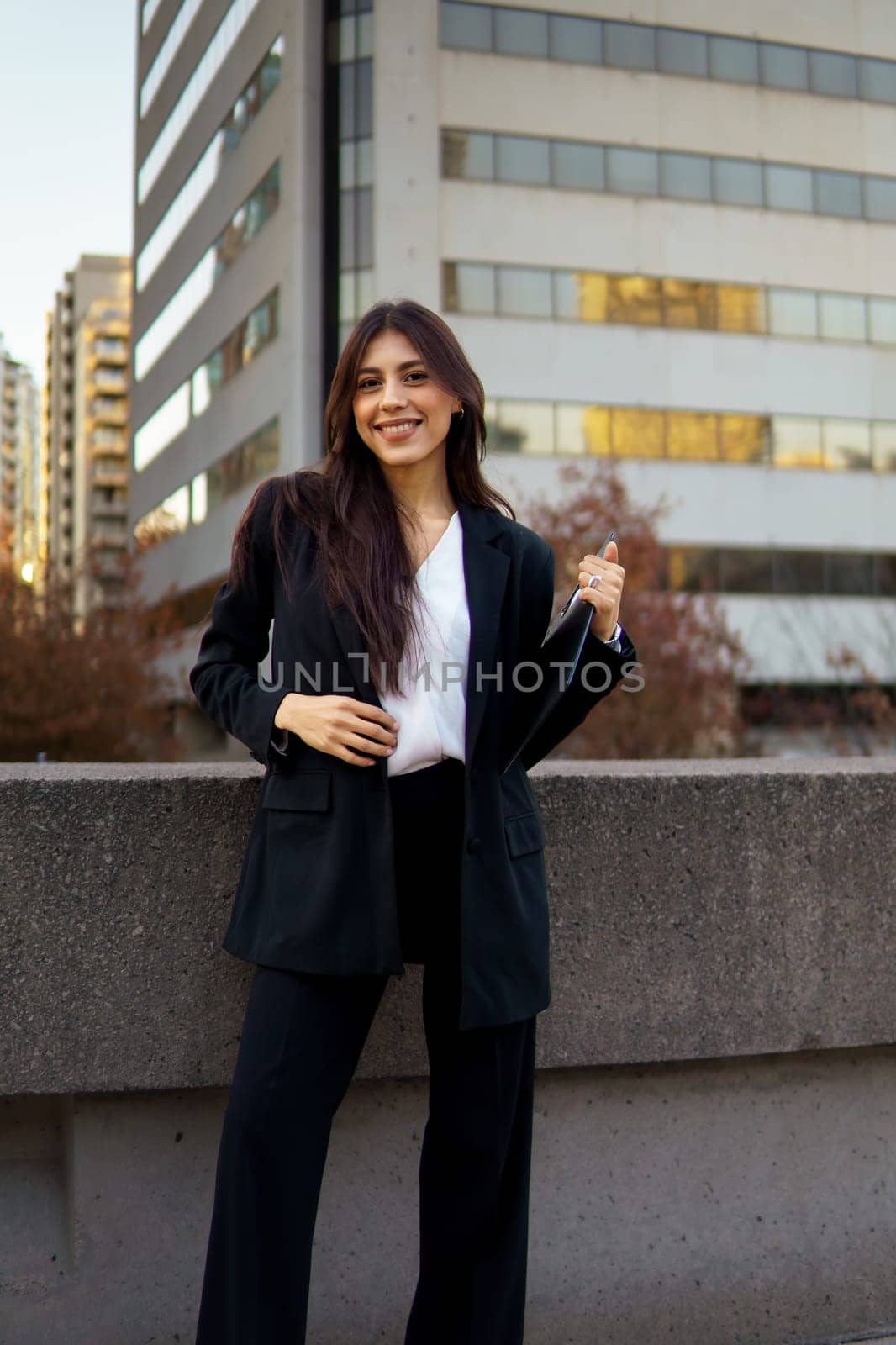 Professional businesswoman standing on a sidewalk in front of a modern building with autumn foliage.