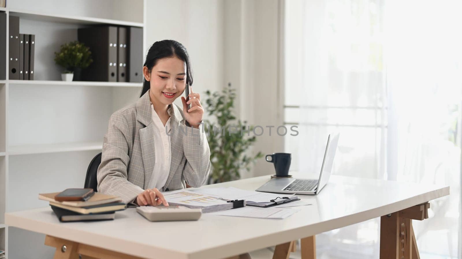 Smiling female accountant talking on mobile phone and checking financial document at office.