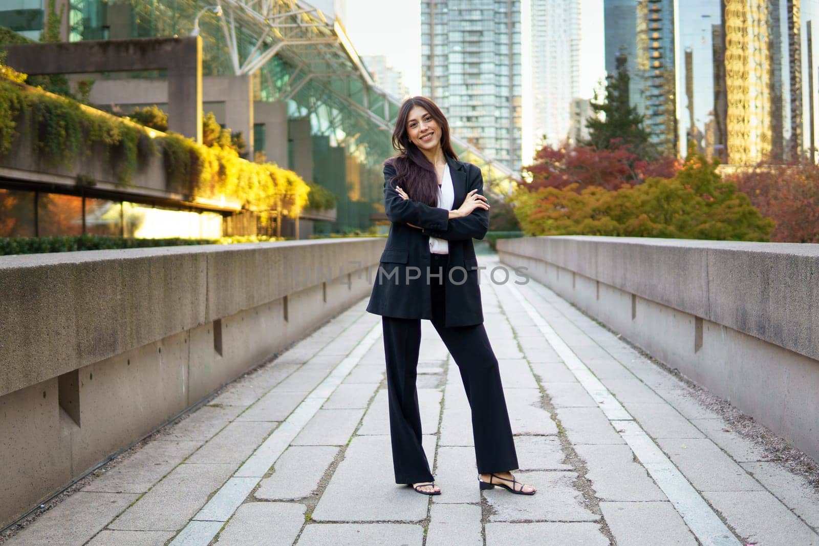Confident young businesswoman in formal suit standing with arms crossed in the middle of an urban pathway.