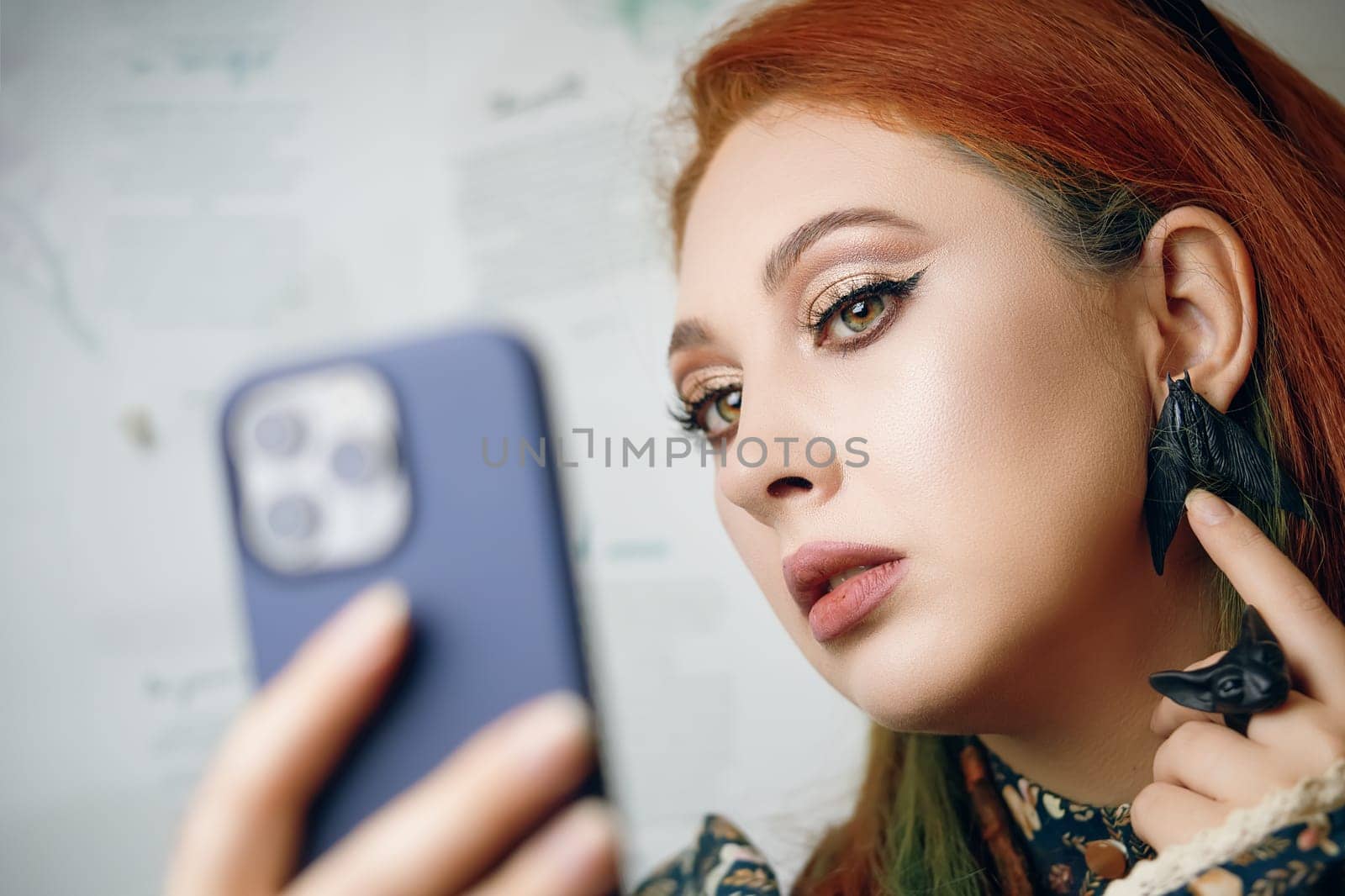 Red-haired woman takes selfie on front camera of modern smartphone. Female blogger films herself for fashion and makeup blog. Cute girl points her finger at her butterfly earrings. Gothic aesthetic.