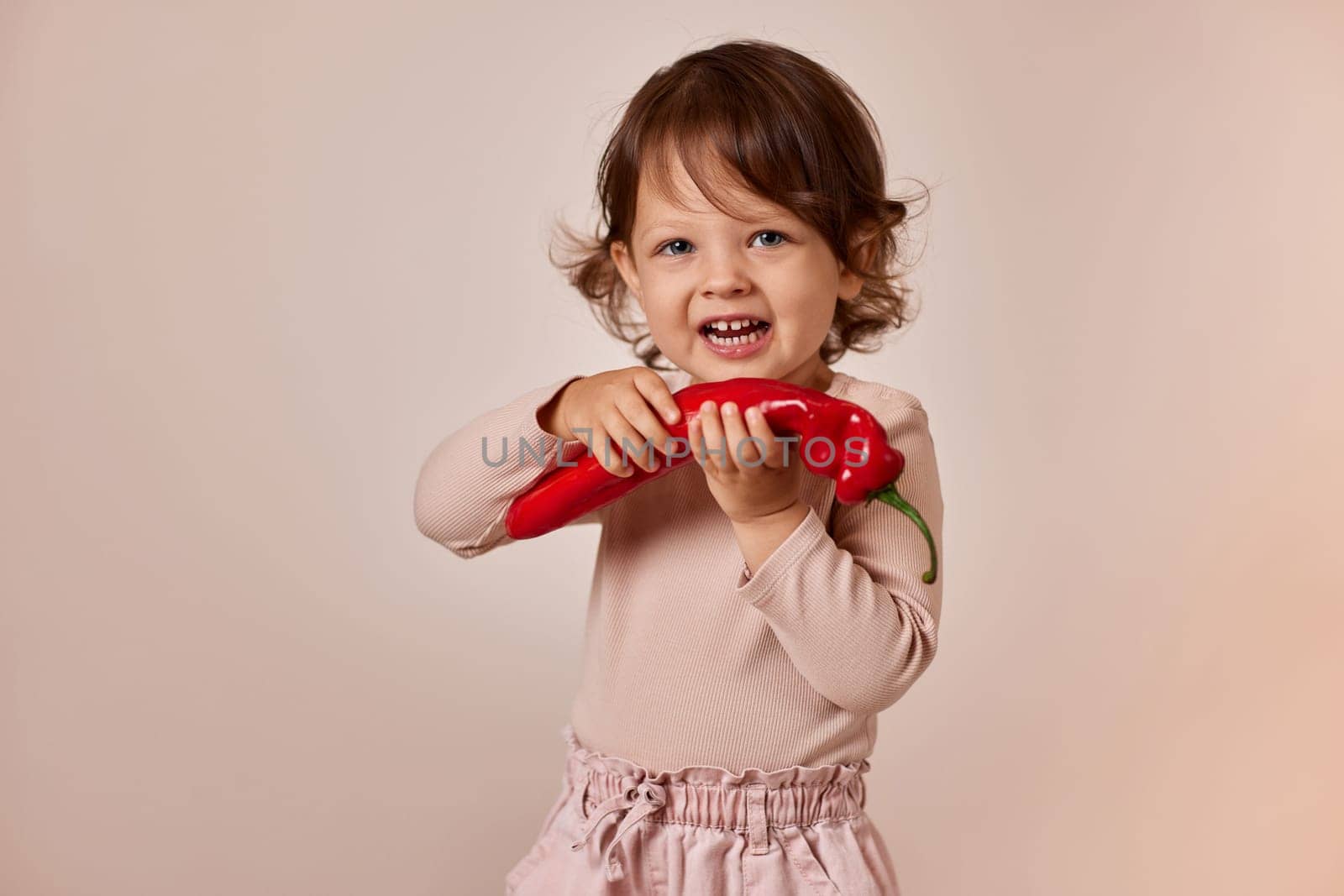 very excited happy child girl holding fresh red pepper on beige background. healthy baby food. kid loves vegetables