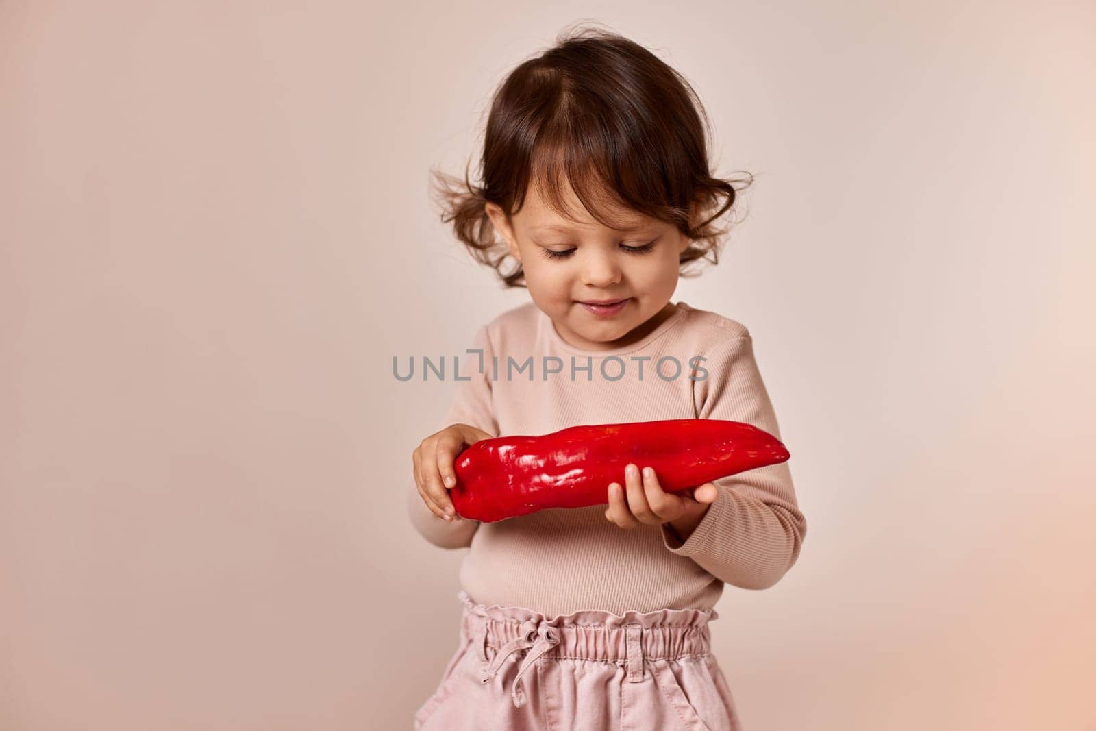 happy child girl holding fresh red pepper on beige background. healthy baby food. kid loves vegetables
