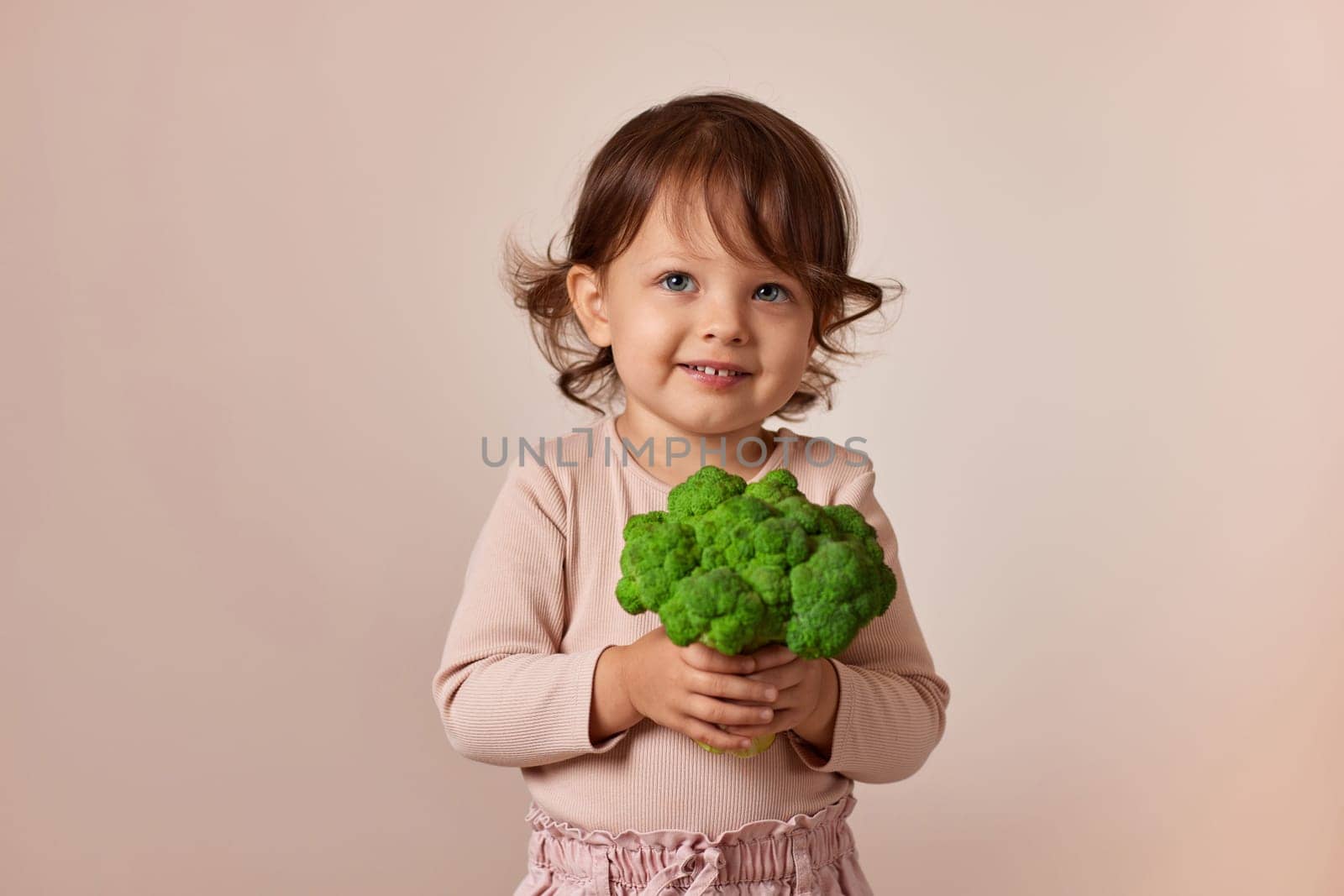 smiling child girl holding fresh green raw broccoli on beige background. healthy baby food. kid loves vegetables