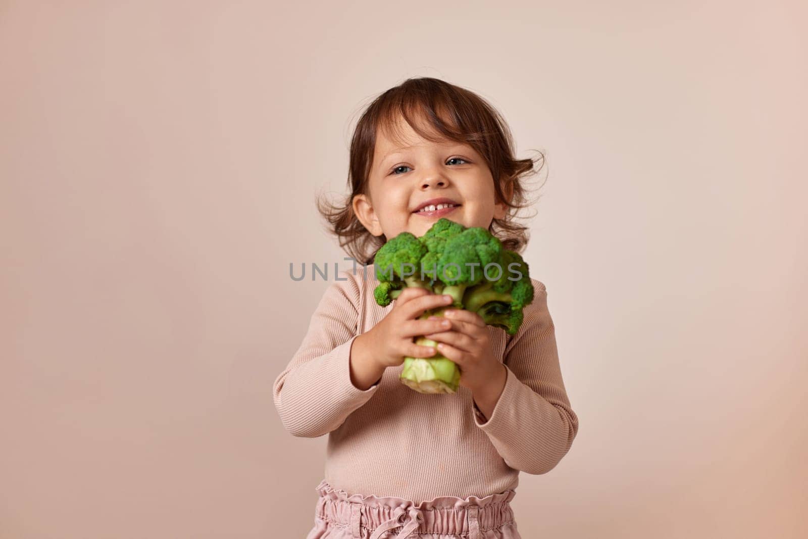 cute child girl holding fresh green raw broccoli on beige background. healthy baby food. kid loves vegetables