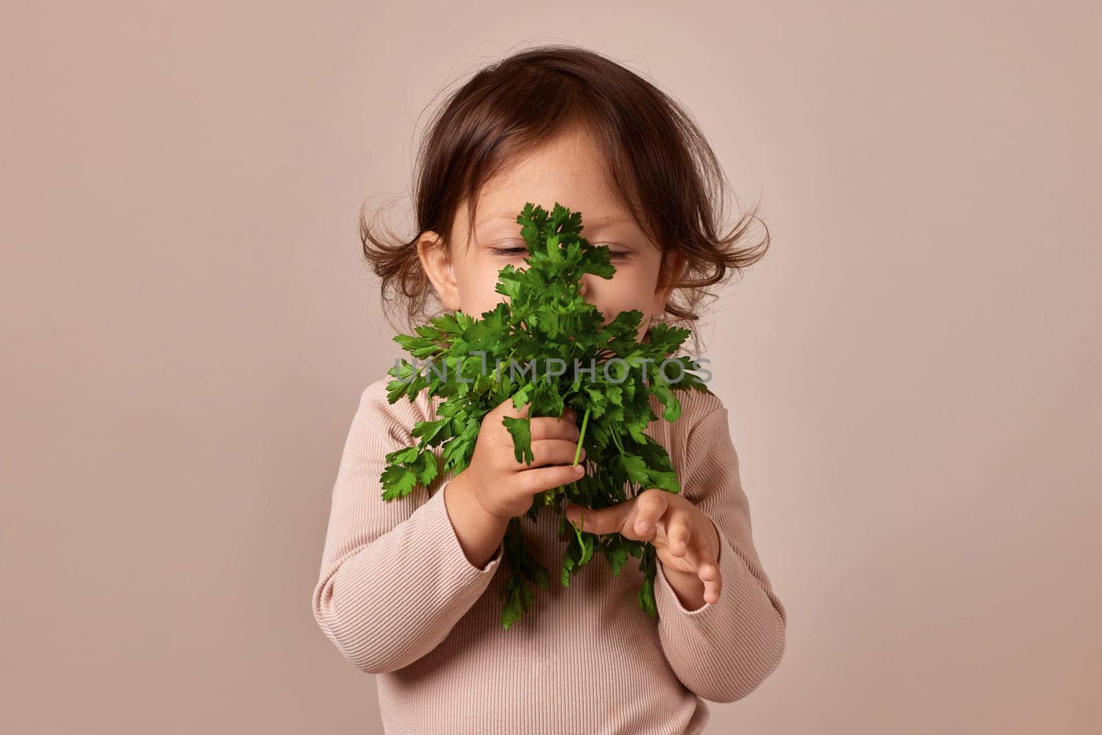 little child girl holding fresh green parsley on beige background. healthy baby food.