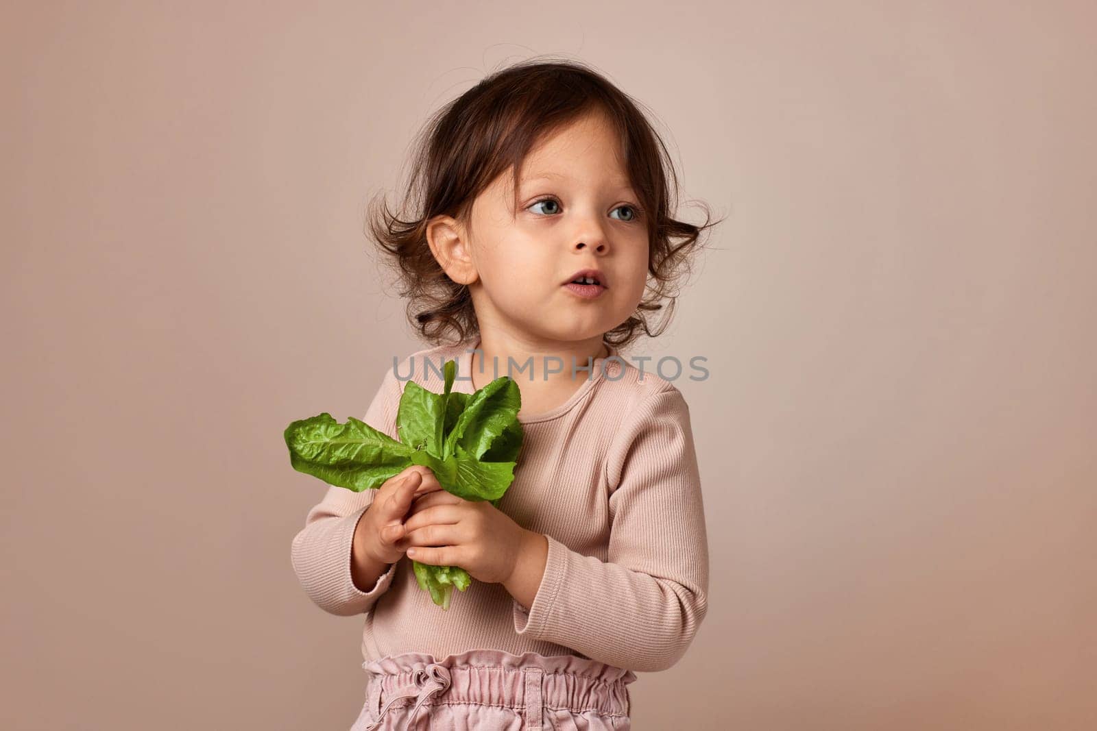 surprised child girl holding fresh green salad on beige background. healthy baby food.