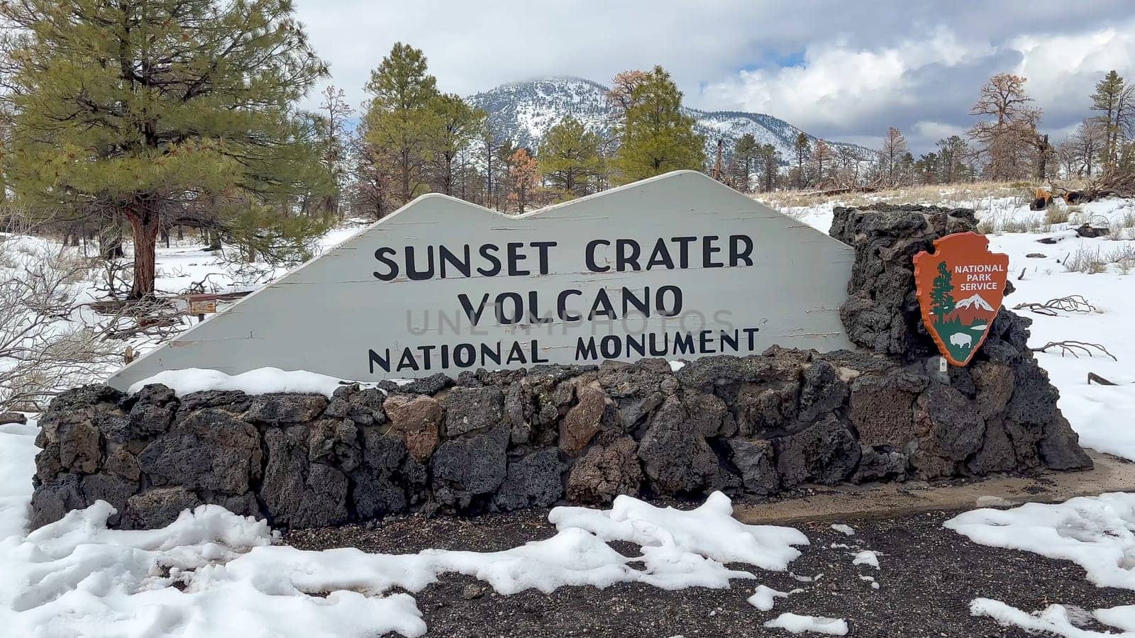 Flagstaff, Arizona, USA - 03.19.2024 - Sunset Crater Volcano National Monument entrance sign on a snowy winter day. The sign is made of wood and stone, and it is surrounded by snow-covered ground.