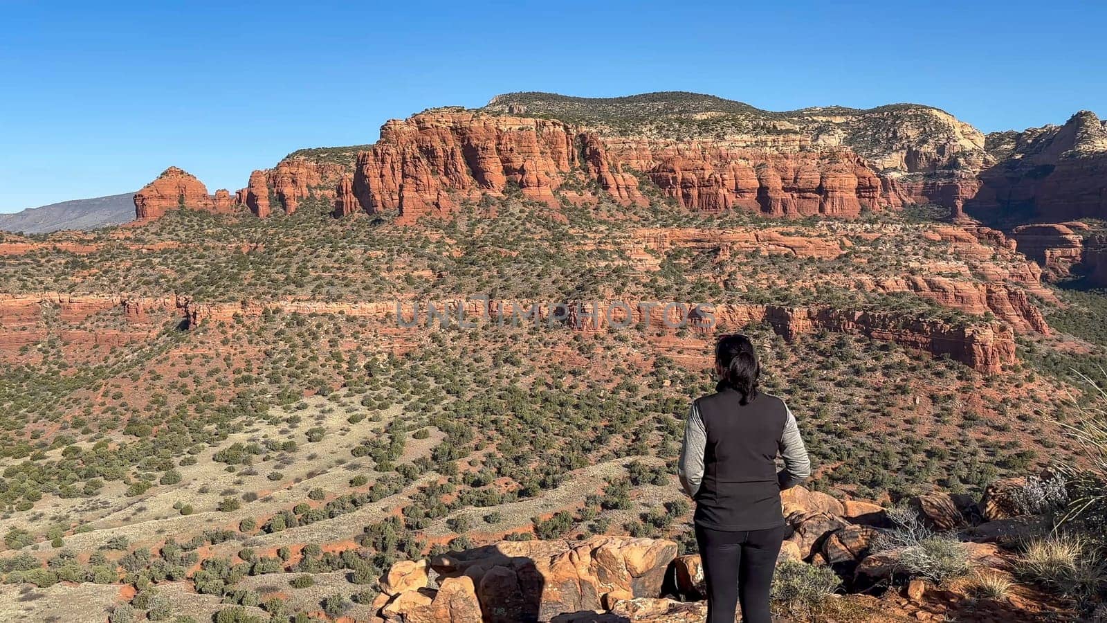 A female hiker stands in awe of the breathtaking panoramic views of Sedona, Arizona's iconic red rock mountains. The vast desert landscape of mesas and buttes stretches out before her - USA