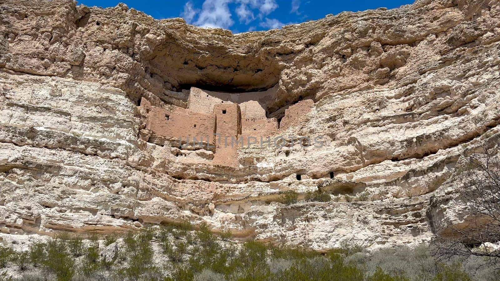 A low-angle view of Montezuma Castle National Monument in Arizona. The ancient Sinagua cliff dwellings are built high up into a limestone alcove. White clouds dot the blue sky above - USA