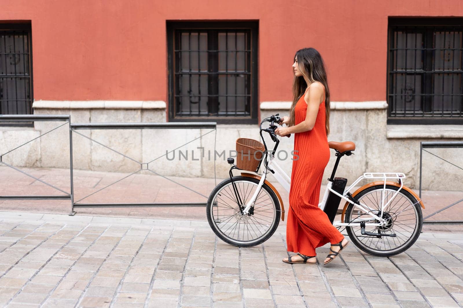 A fashionable woman in a vibrant red dress strolls beside her chic ebike along a colorful wall, embodying an elegant urban lifestyle that showcases her unique style and charisma