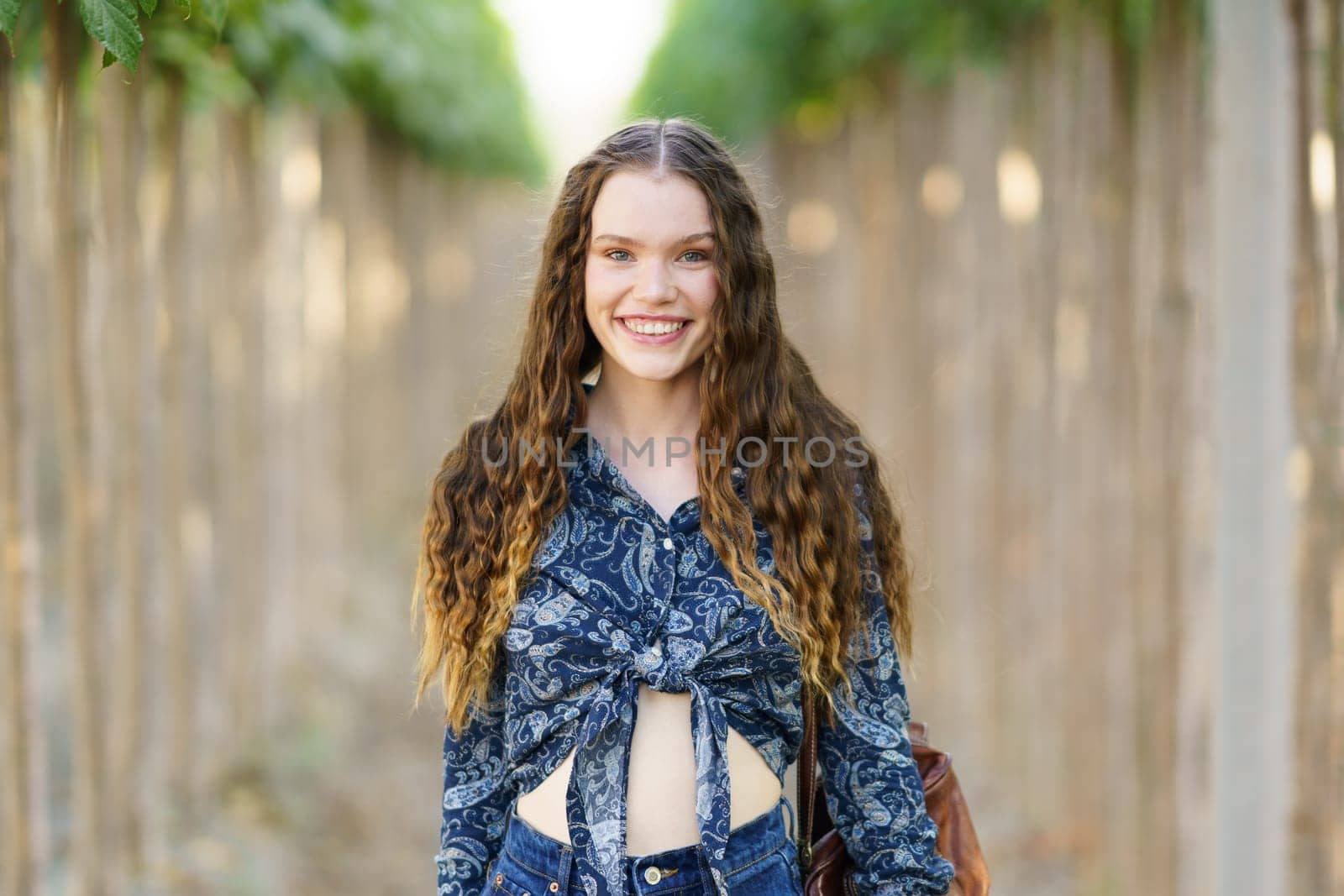 A cheerful young woman with curly hair stands confidently at a charming pathway, which is beautifully lined with an array of vibrant trees, capturing a truly magical moment of natural beauty
