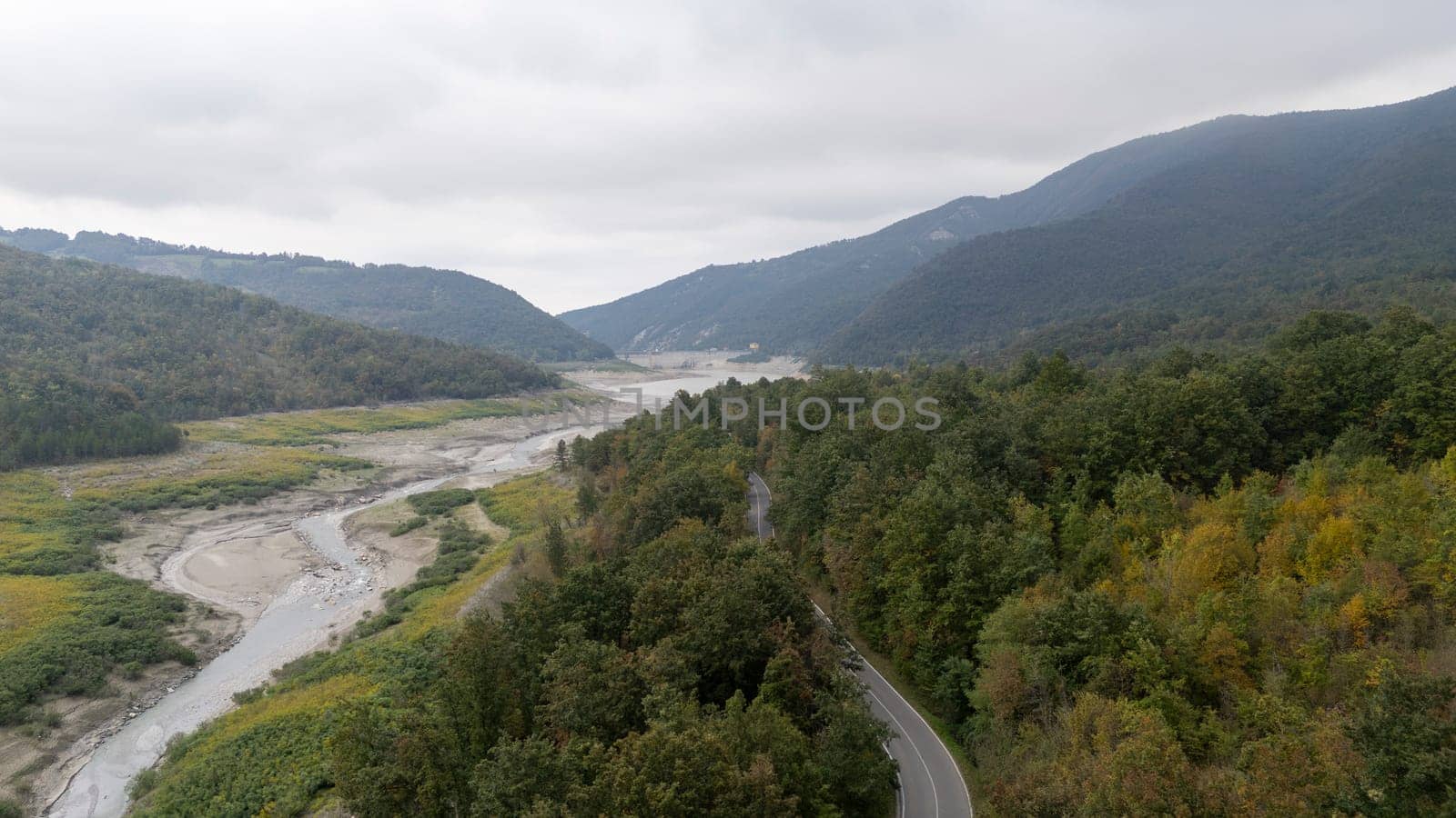 Asphalt road is winding through a lush green forest towards a lake with a very low water level. Lago di Mignano, Val Tolla, PC, Italy