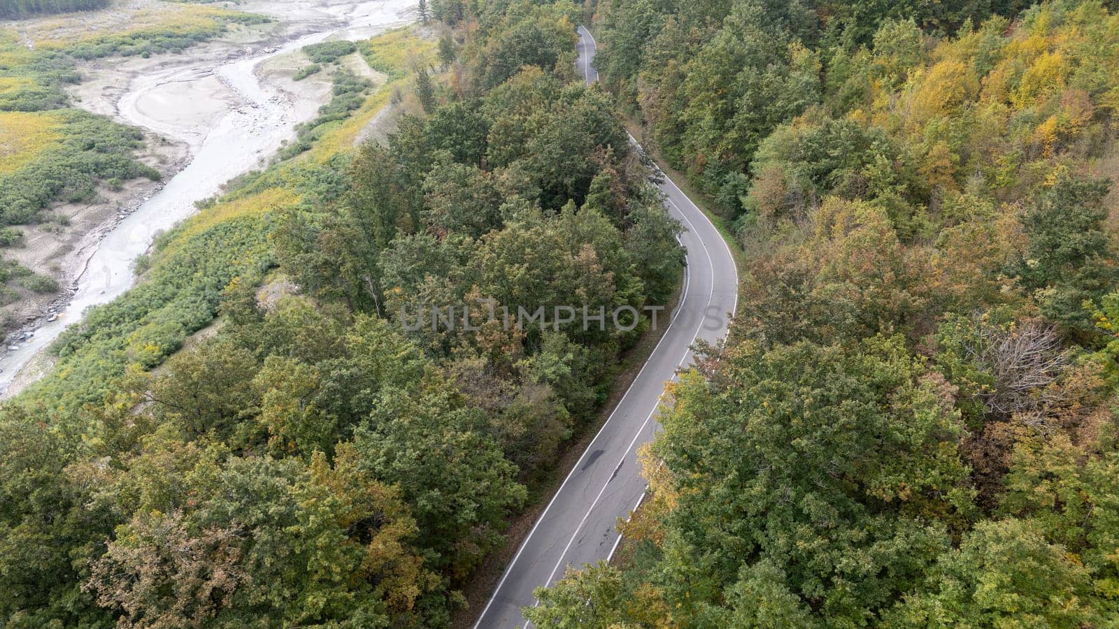 Aerial view of a winding road going through a forest next to a river in autumn. Lago di Mignano, Val Tolla, PC, Italy