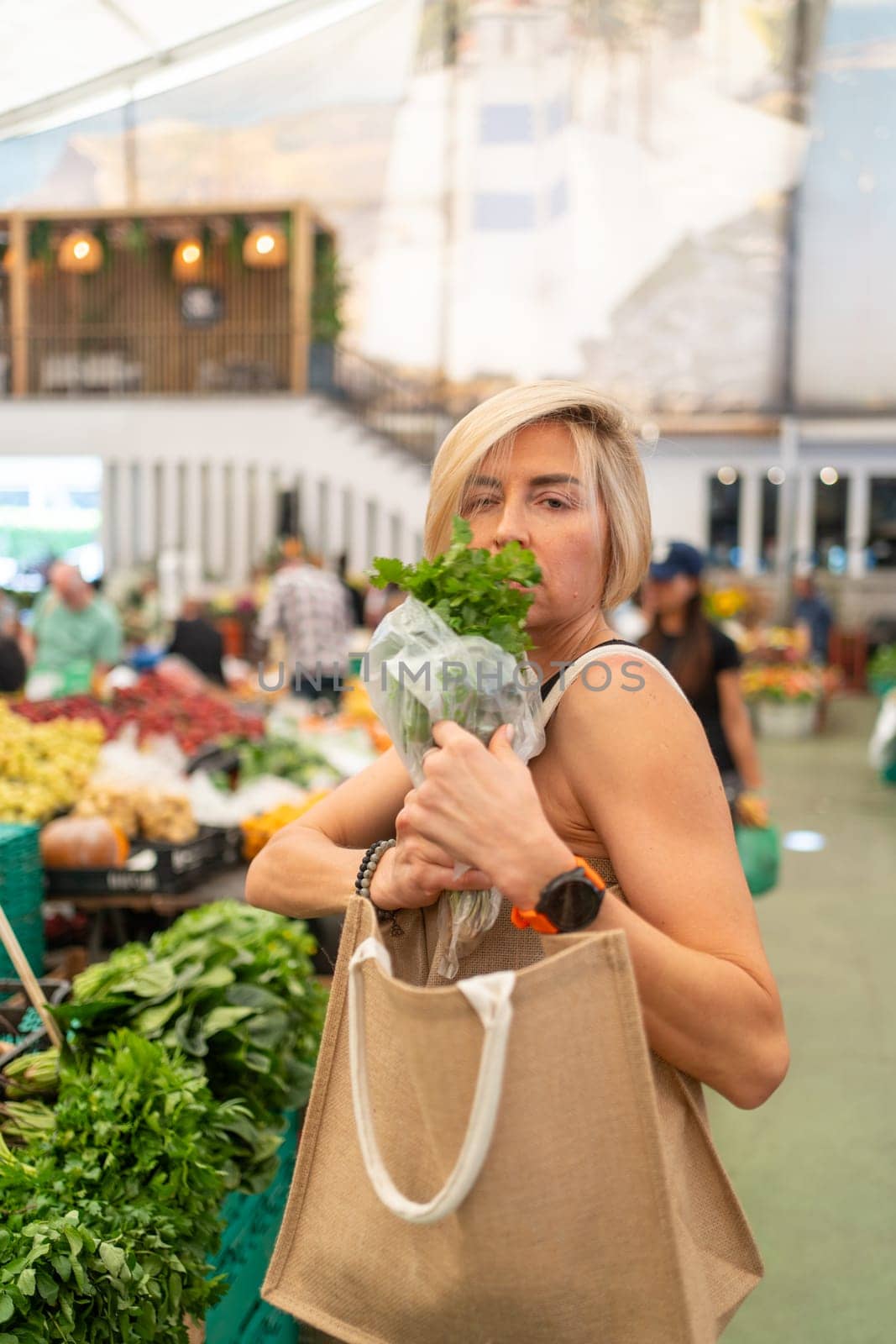 Woman holding fresh basil leaves at supermarket during daytime. Side view of Caucasian female looking at camera while shopping for groceries in city.