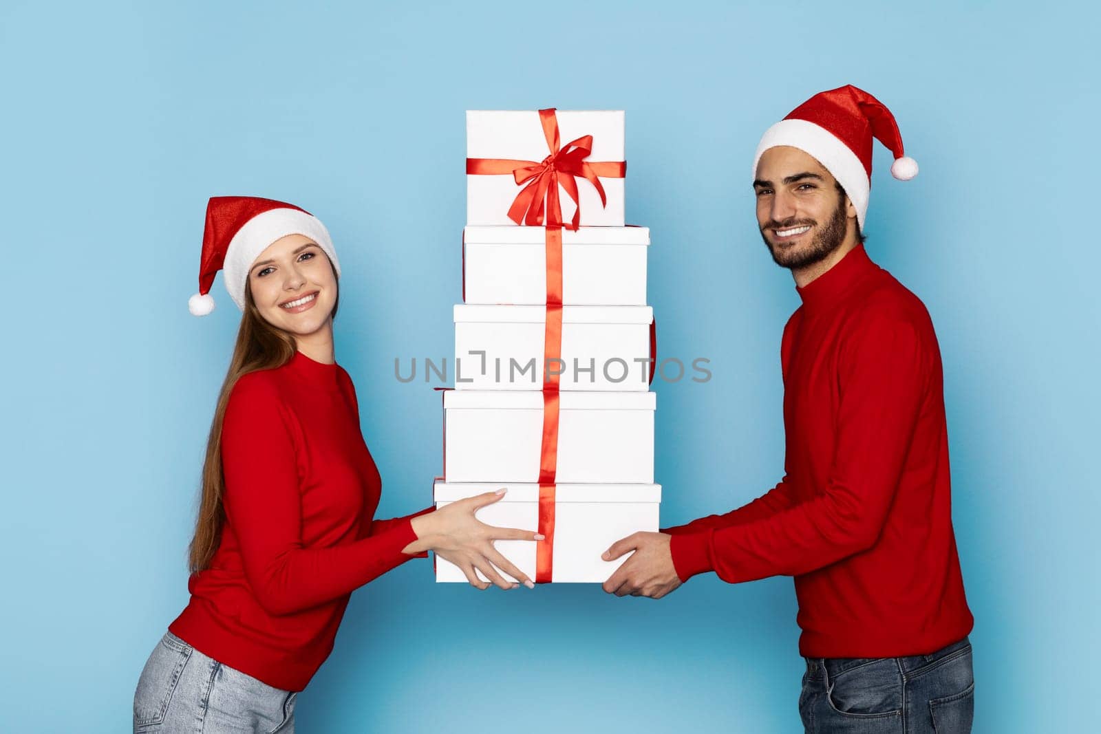 A happy couple wearing red sweaters and Santa hats joyfully holds a stack of decorated Christmas gifts. The blue backdrop enhances the festive spirit.
