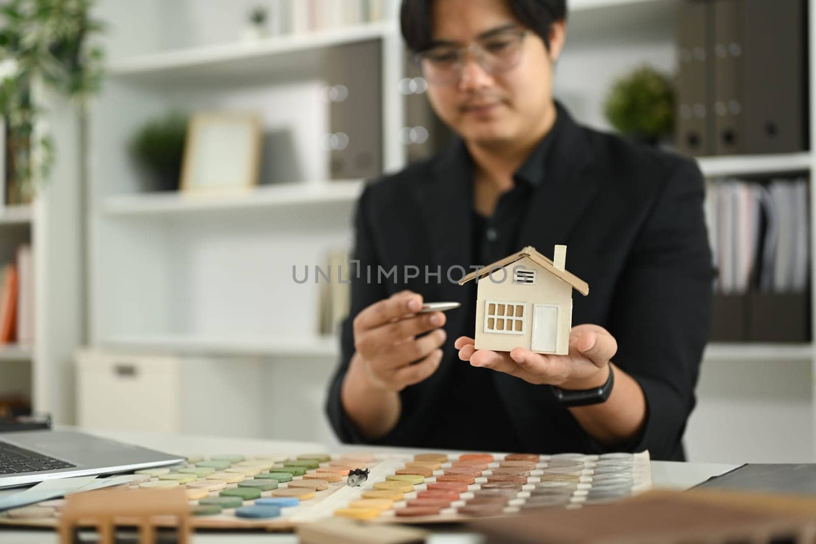 Man holding a small model house while reviewing details of the model at desk with color samples and material swatches.