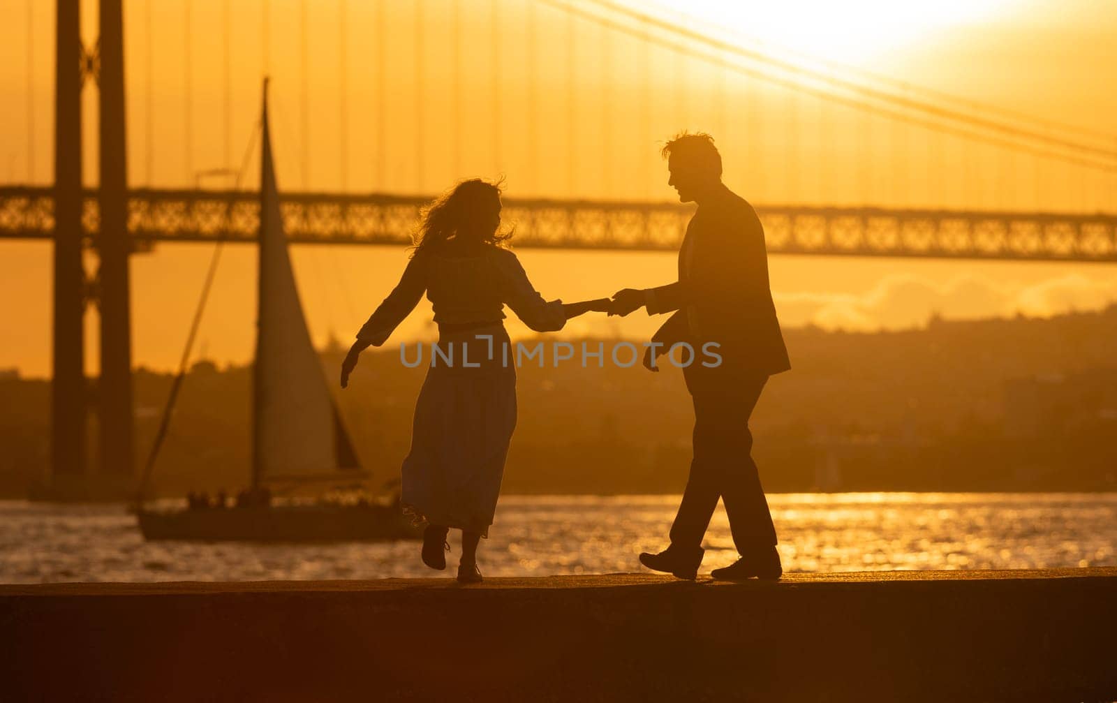 Silhouette of a couple holding hands and dancing by the river with a bridge and sunset in the background