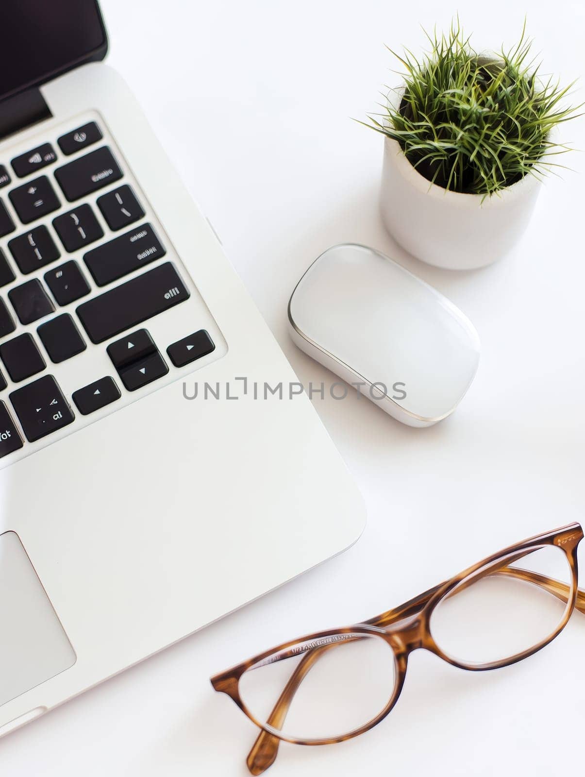 A clean, white desktop workspace featuring a laptop, wireless mouse, eyeglasses, and a small potted plant, embodying modern minimalist office design with copy space.