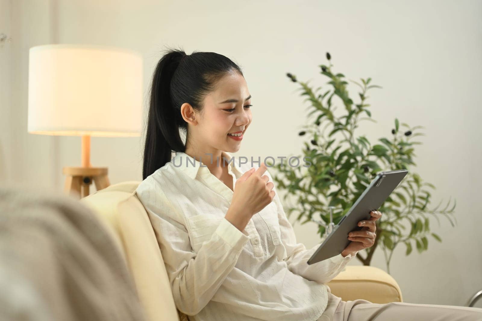 Smiling young woman wearing casual dressed using digital tablet on sofa at home.