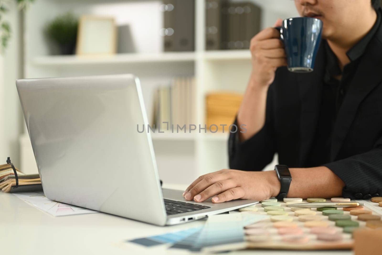 Male architect sipping coffee while working on a laptop at desk with model buildings and material samples.