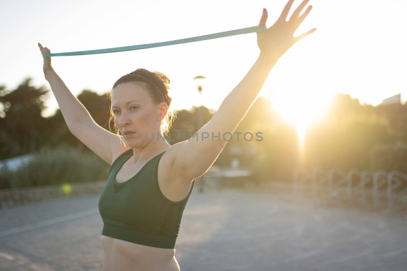 Athletic woman is using a resistance band while working out outside at sunset