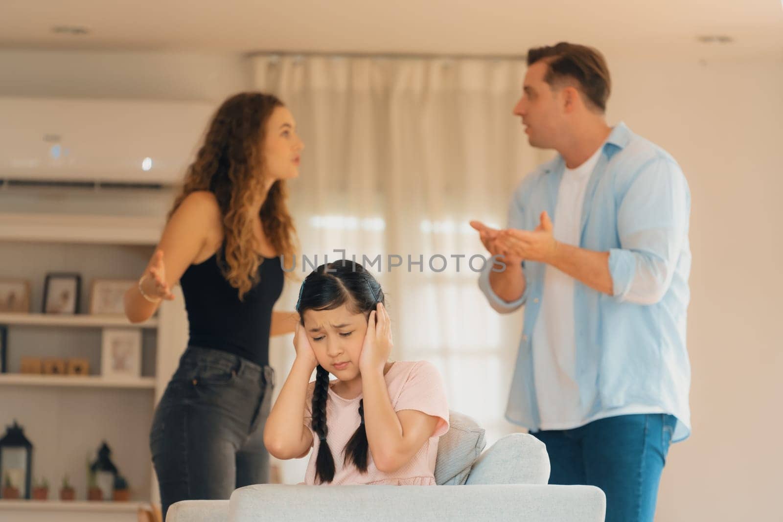 Annoyed and unhappy young girl sitting on sofa trapped in middle of tension by her parent argument in living room. Unhealthy domestic lifestyle and traumatic childhood develop to depression.Synchronos