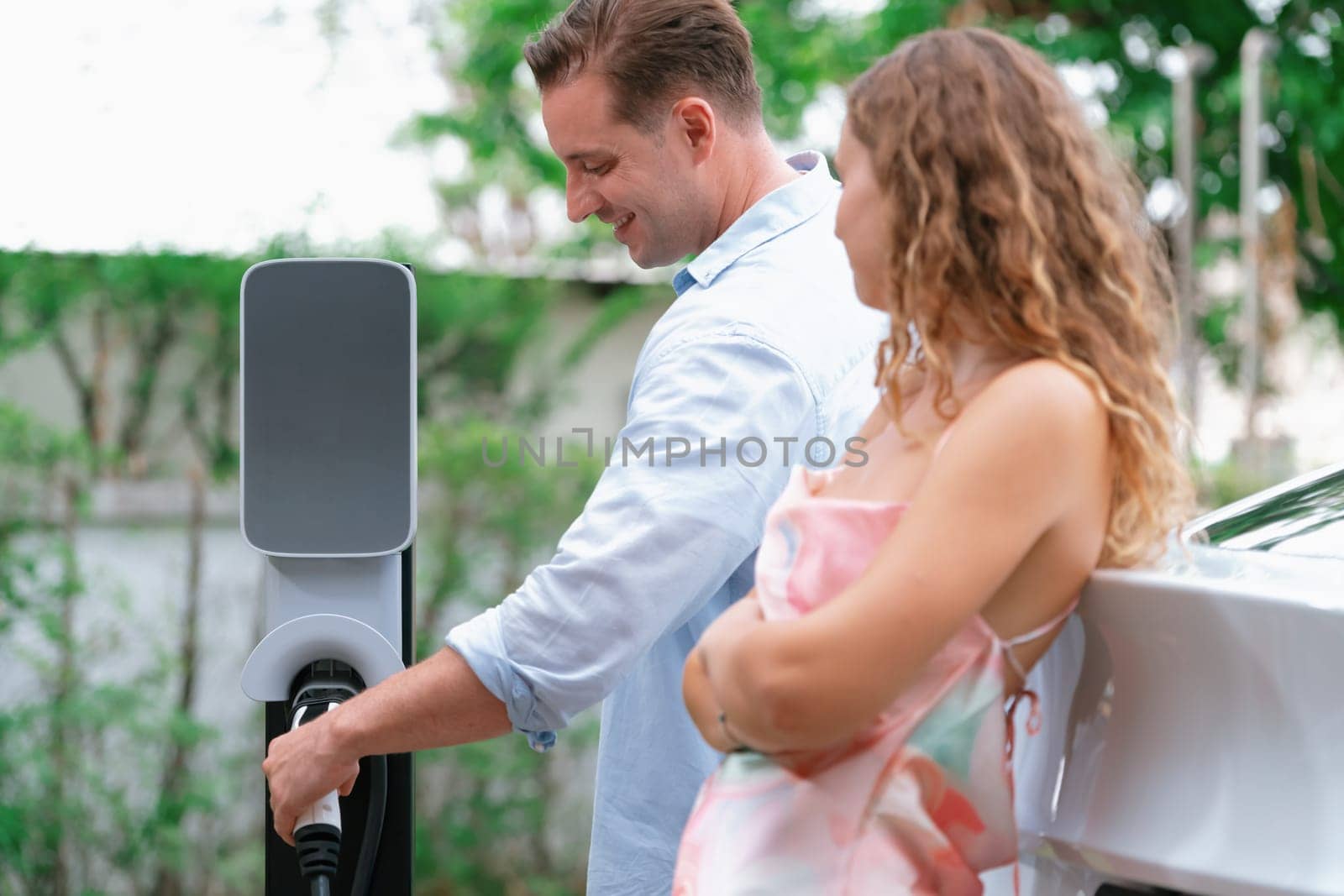 Happy and lovely couple with eco-friendly conscious recharging electric vehicle from EV charging station. EV car technology utilized as alternative transportation for future sustainability. Synchronos