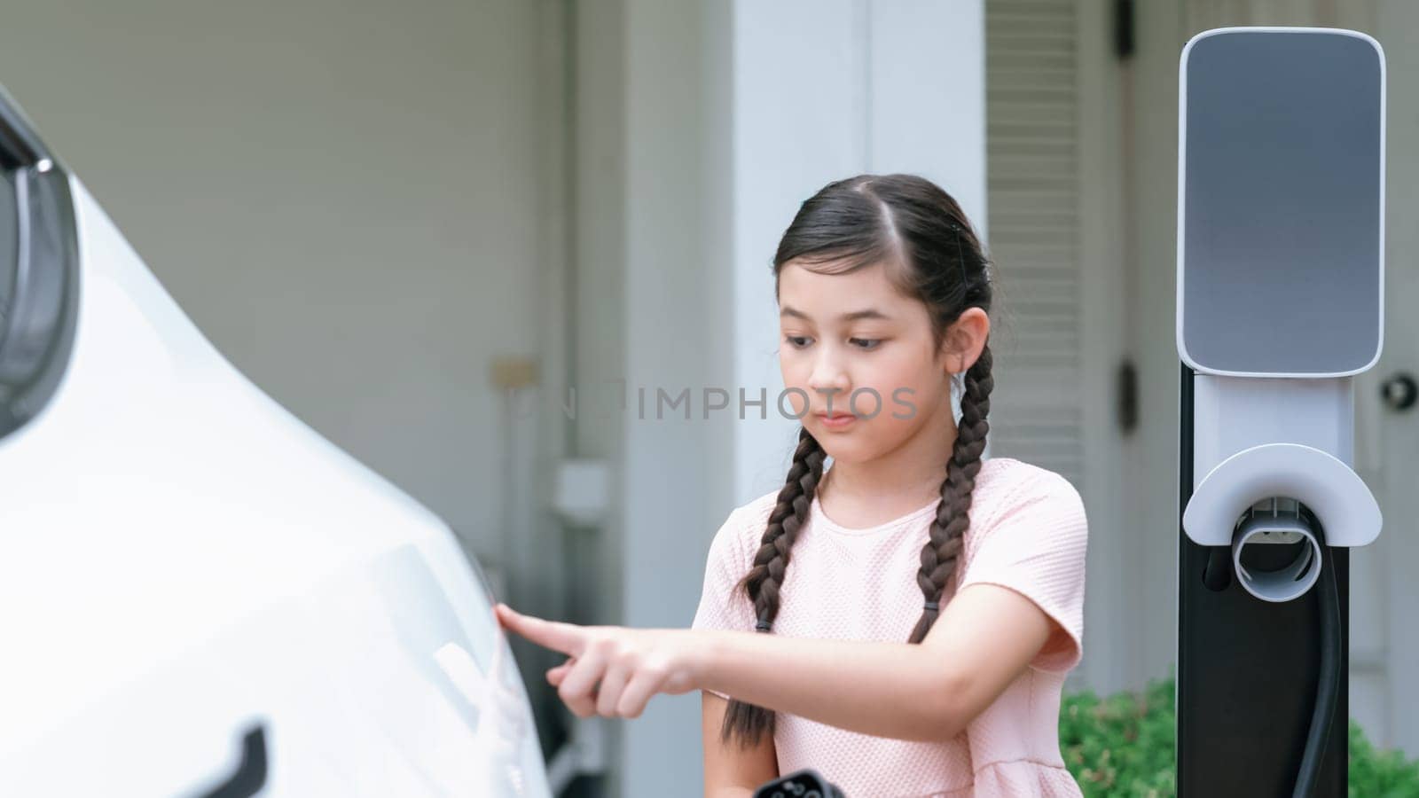 Happy little young girl learn about eco-friendly and energy sustainability as she recharge electric vehicle from home EV charging station. EV car and sustainable future generation. Panorama Synchronos