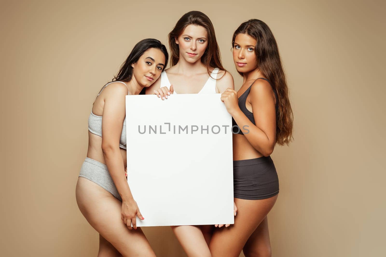 Serious millennial arab, latin, caucasian women in underwear enjoy self love hold banner with copy space, isolated on beige background, studio. Beauty care, motivation, lifestyle