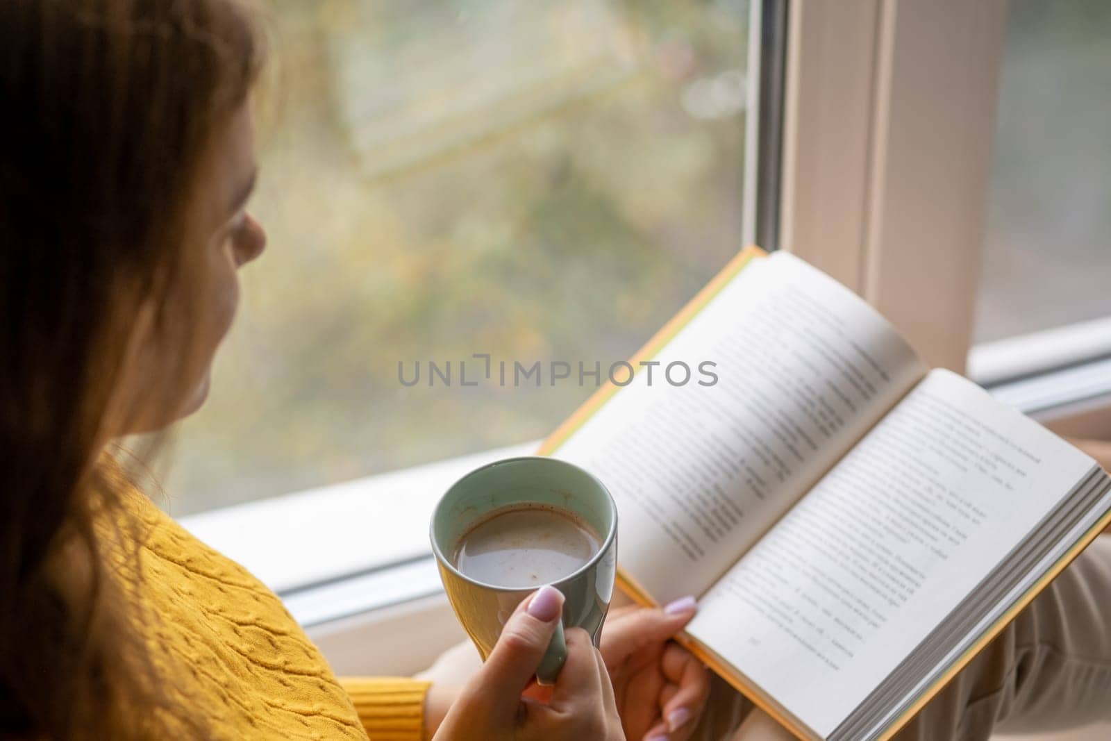 Young beautiful woman near window yellow knitted sweater read book by AnatoliiFoto