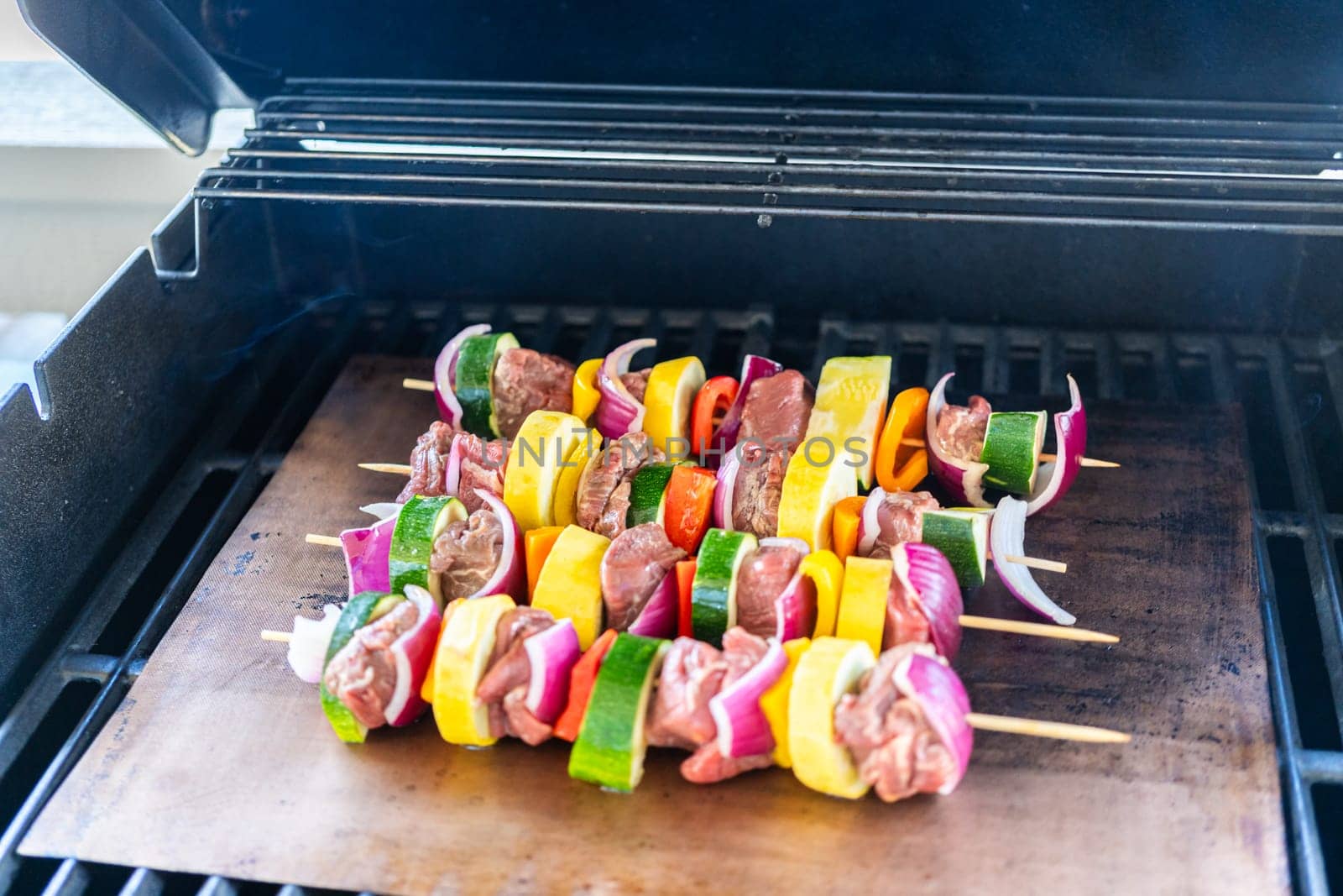 Skewered beef and fresh veggies sizzle on a copper grill mat over a gas outdoor grill.