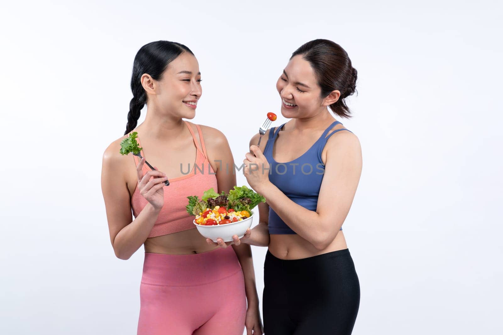 Two young sporty Asian women in sportswear holding salad bowl fill with fruit and vegetable. Natural youthful and fit body lifestyle people with balance nutrition on isolated background. Vigorous