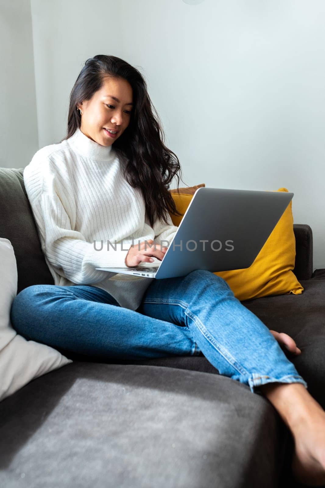 Young Asian woman using laptop at home sitting on the couch, relaxing. Vertical image. by Hoverstock
