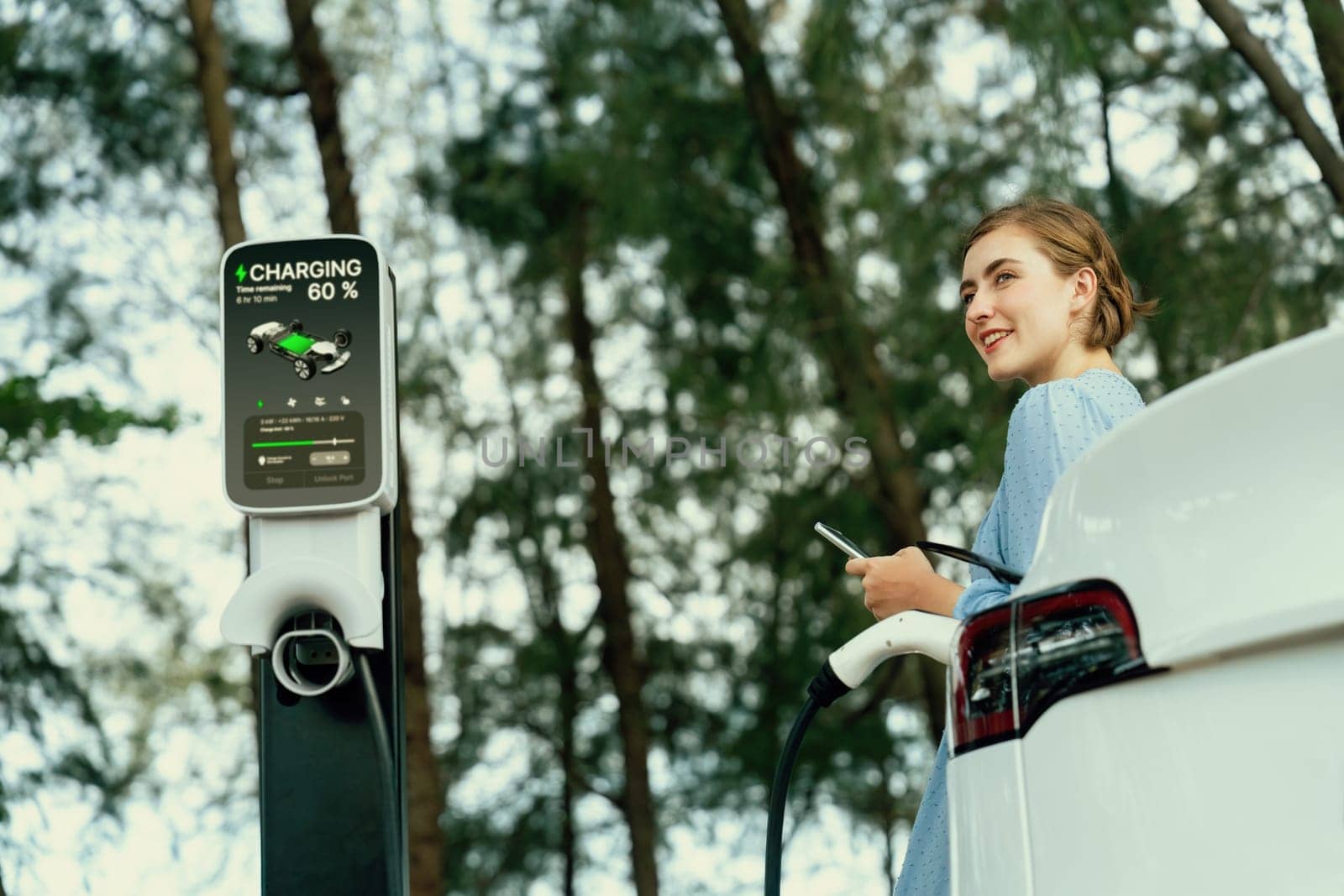 Holiday road trip vacation by the green countryside nature with beautiful young woman recharging electric vehicle with alternative energy. Environmental friendly travel wit EV car. Perpetual