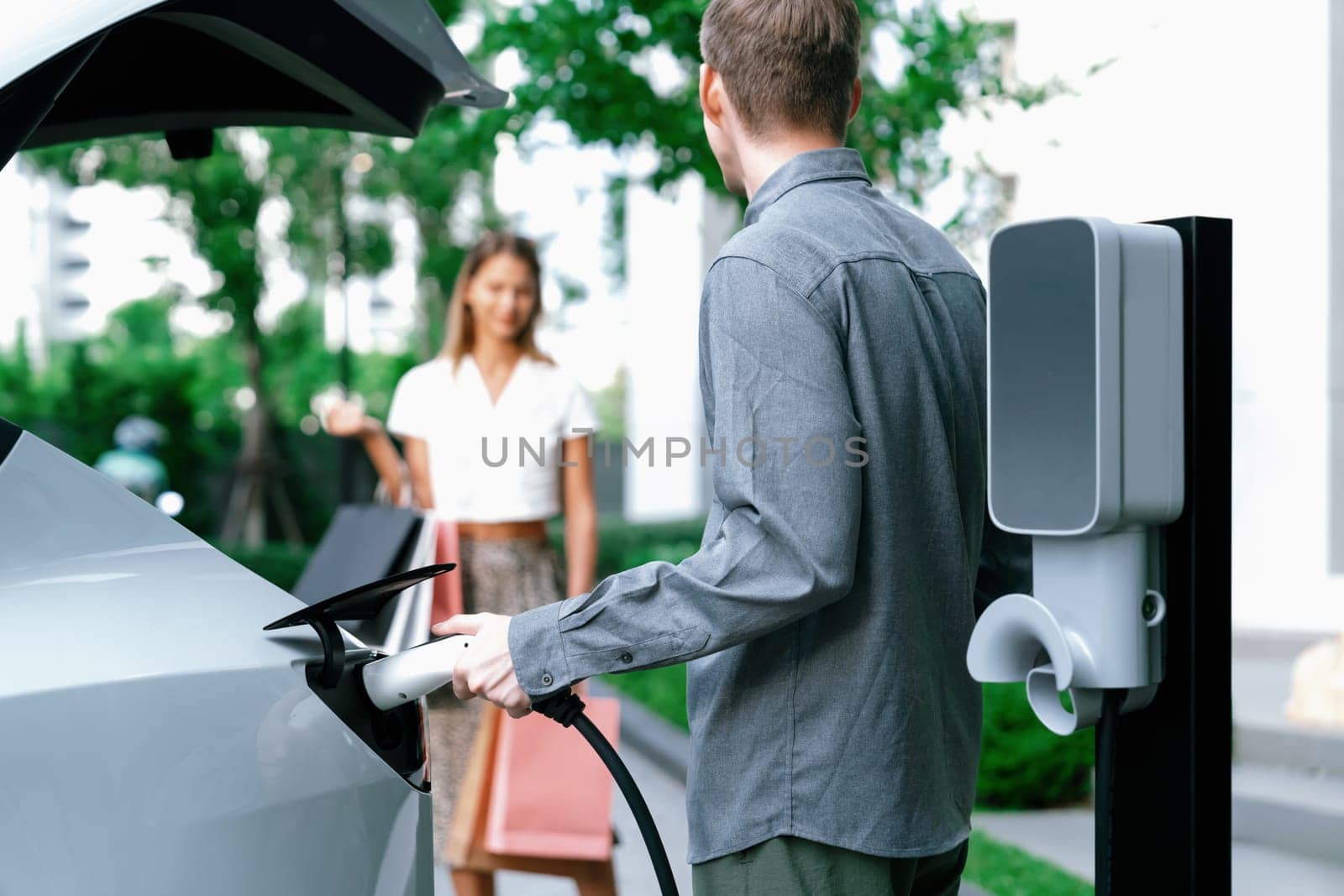 Young couple travel with EV electric car charging in green sustainable city outdoor garden in summer shows urban sustainability lifestyle by green clean rechargeable energy of electric vehicle innards