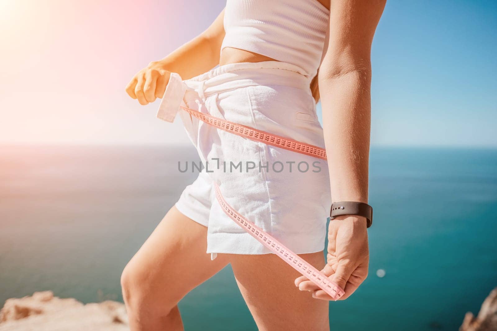 a young and slender woman in shorts holds in her hand a measuring yellow tape on her sexy thighs on the beach. concept of detox and diet